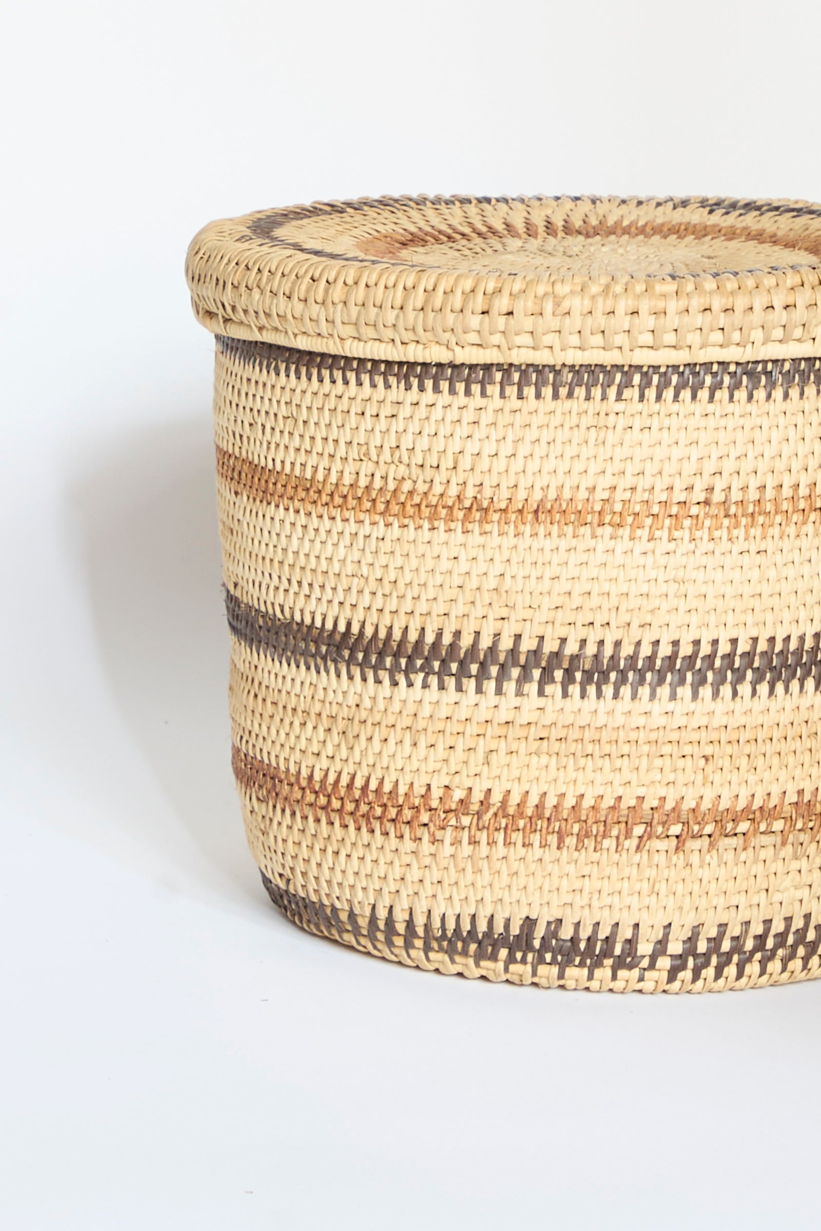 A round Motorohina Basket with Yanomami Stripe Painting Mixed by Incausa, handwoven with a lid and featuring alternating natural and dark brown horizontal stripes, set against a plain white background.