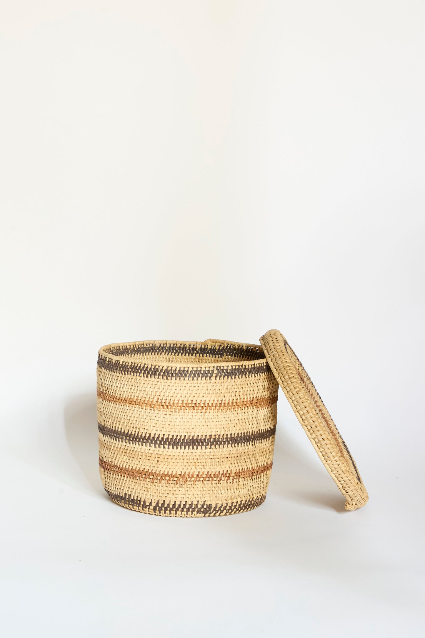 The Motorohina Basket with Yanomami Stripe Painting Mixed by Incausa is displayed with its matching lid set to the side, showcasing its round, woven design and brown-and-beige striped pattern against a plain white background.