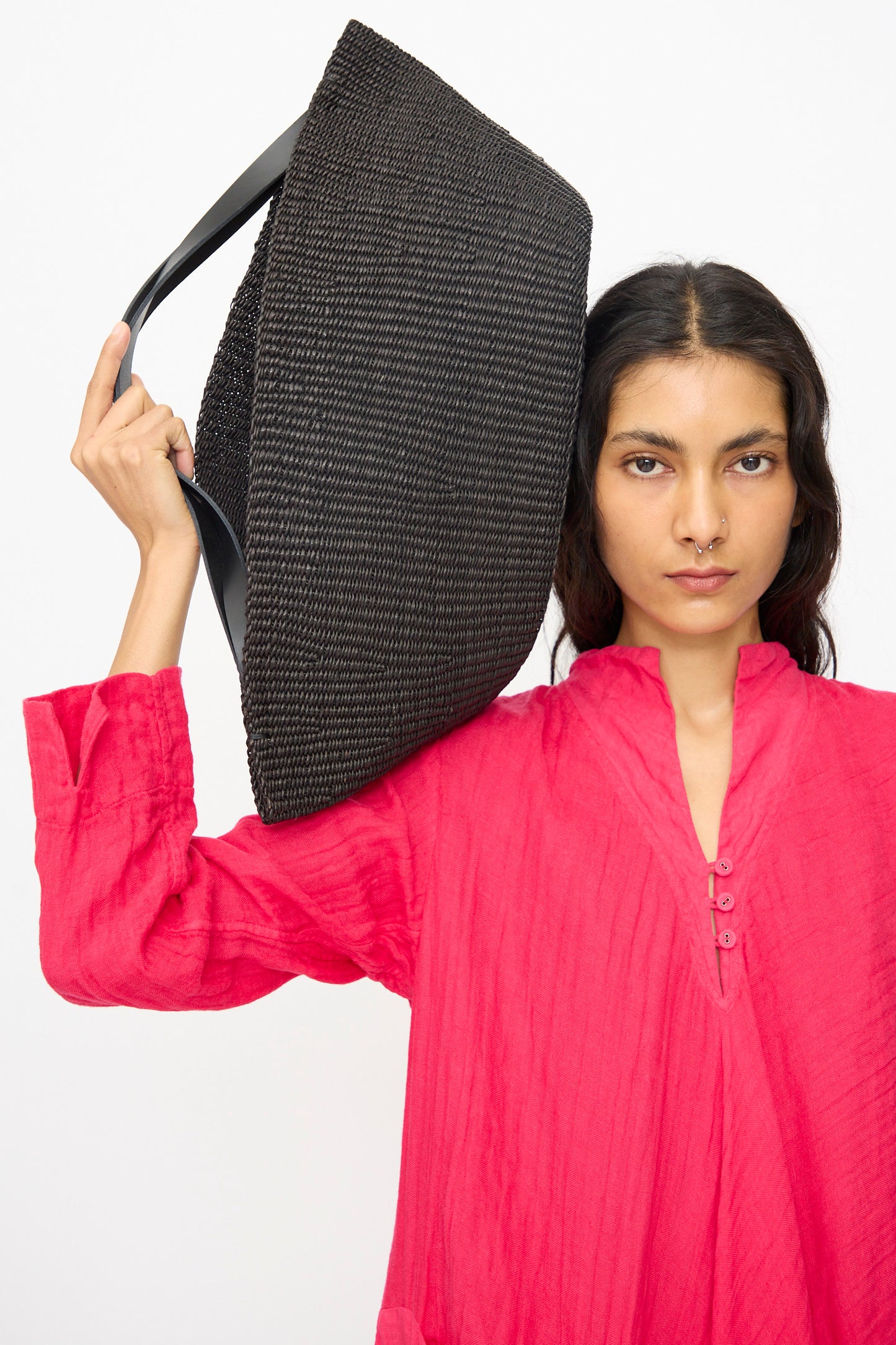 A person in a bright pink long-sleeve top carries the Inès Bressand Large Gousse Bag in Black, handmade in Ghana, on their shoulder against a plain white background.