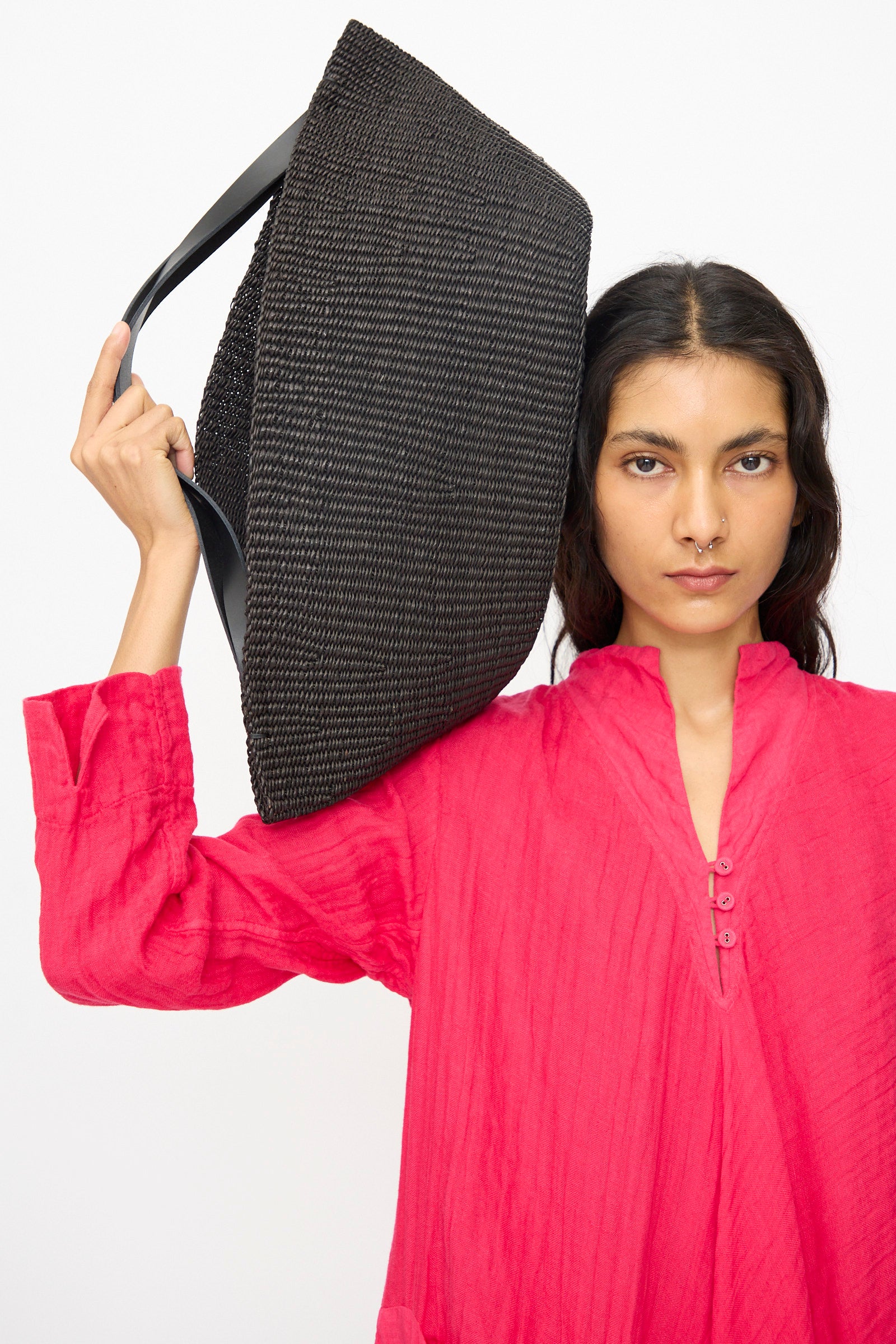 A person in a bright pink long-sleeve top carries the Inès Bressand Large Gousse Bag in Black, handmade in Ghana, on their shoulder against a plain white background.