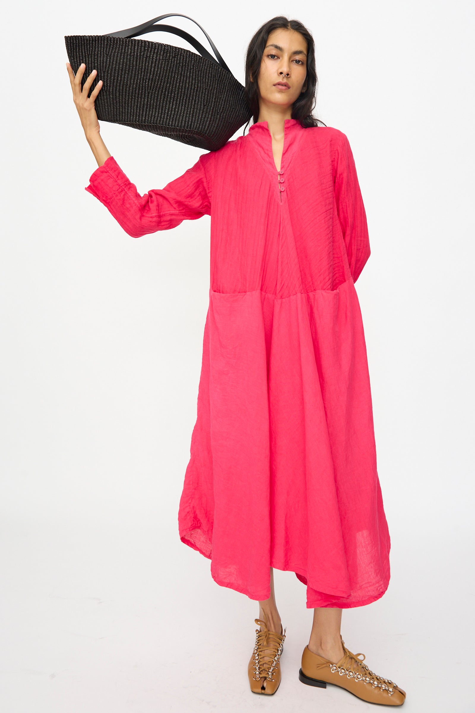A person stands against a plain white background, wearing tan shoes and a loose-fitting bright pink dress, holding the Inès Bressand Large Gousse Bag in Black on their shoulder—an oval handbag handmade in Ghana.