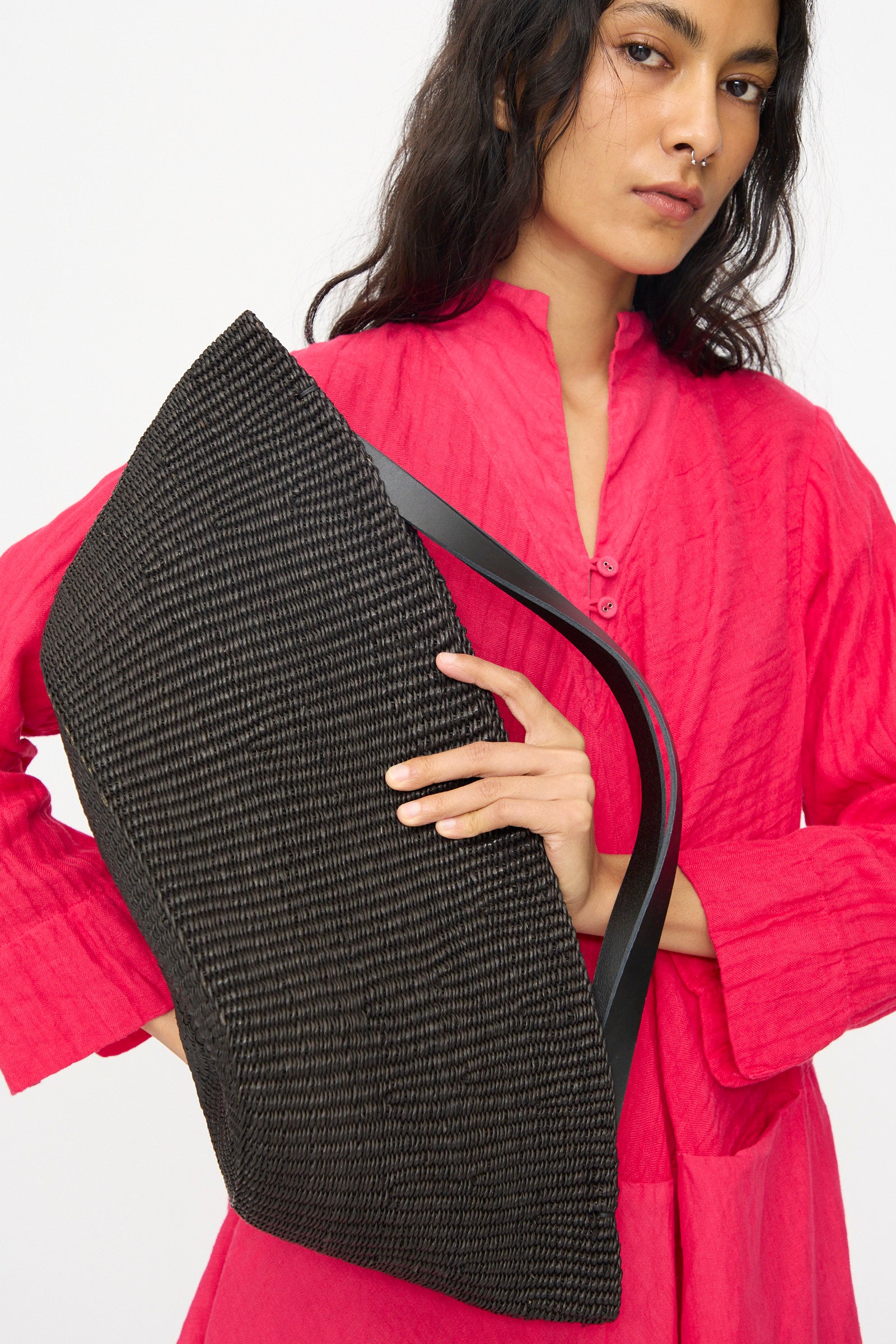 A person in a bright pink long-sleeve dress holds the Large Gousse Bag in Black by Inès Bressand, an elegant oval handbag handmade in Ghana, against a plain light background.