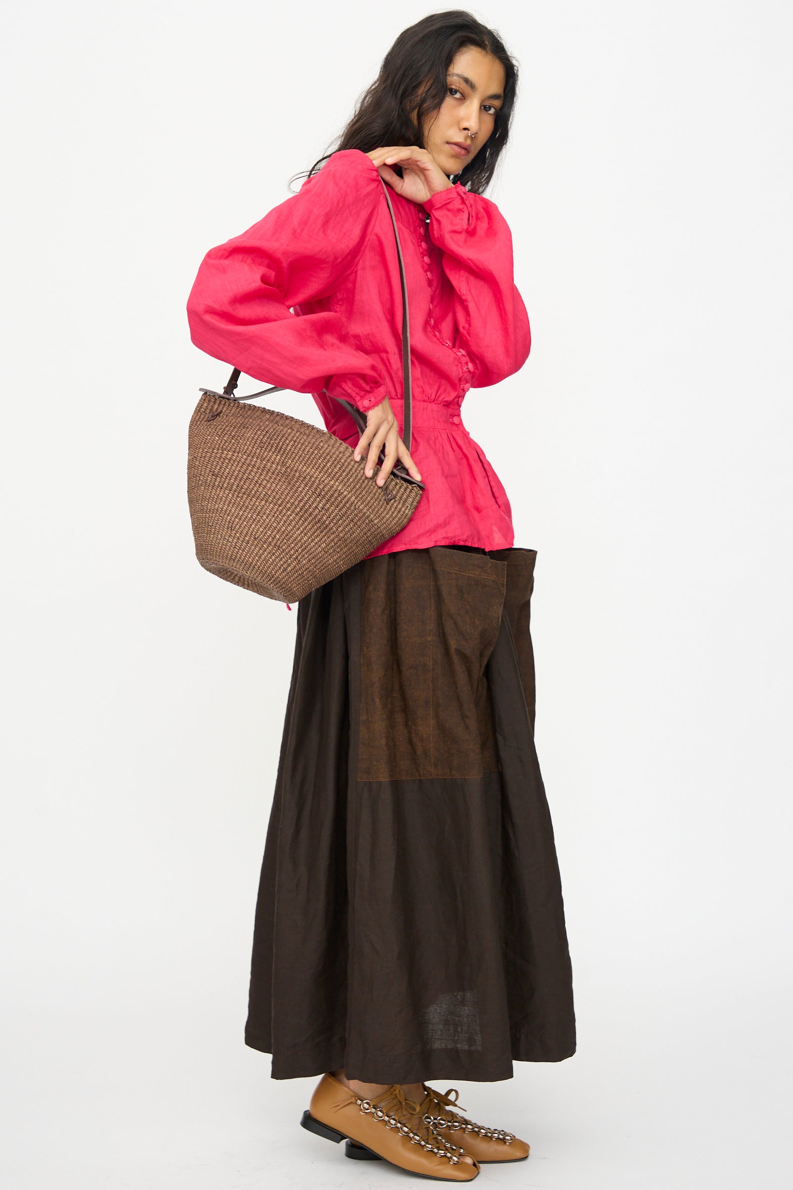 A person stands against a plain white background holding the Inès Bressand Medium Bell Bag in Brown, crafted from elephant grass and handmade in Ghana.