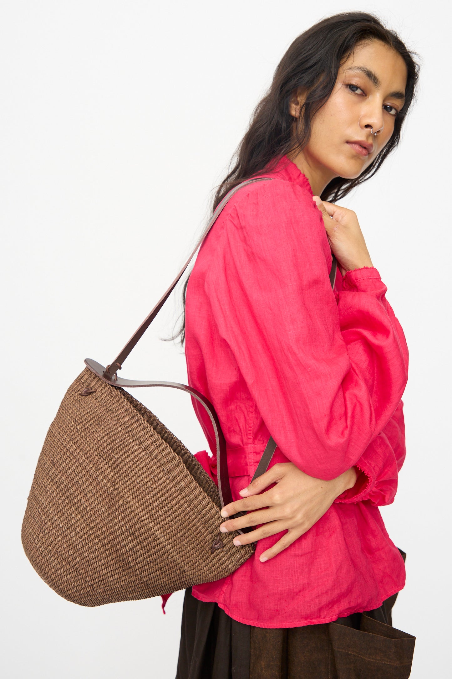 A person wearing a bright pink long-sleeve blouse and a long dark skirt carries the Medium Bell Bag in Brown by Inès Bressand.