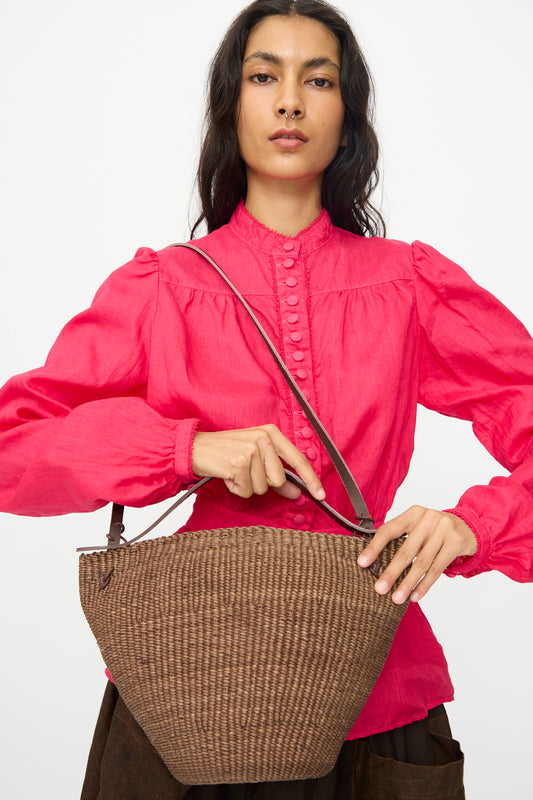 A person in a bright pink blouse holds the Medium Bell Bag in Brown from Inès Bressand—handmade in Ghana from elephant grass with a shoulder strap and vegetable-tanned leather accents—against a plain background.