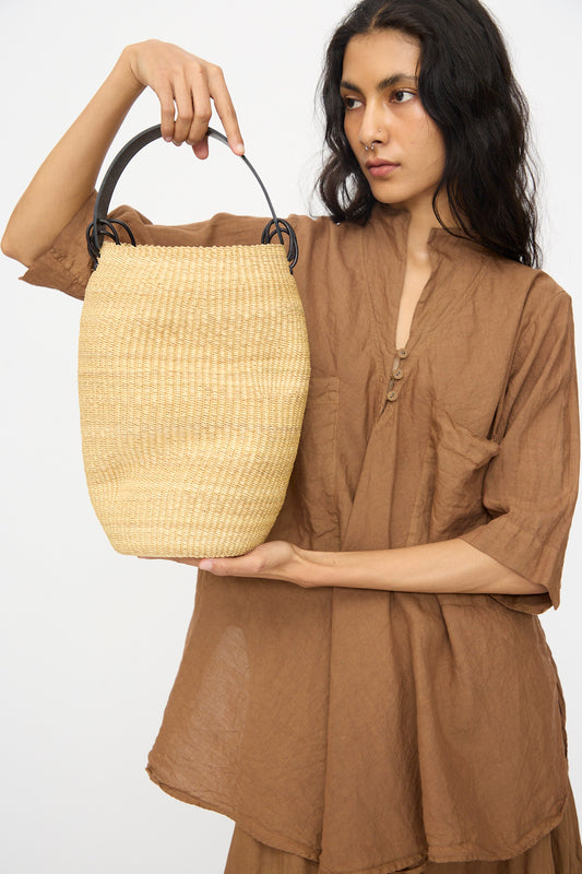 A person in a loose brown outfit holds up the Pot Froissé Bag in Natural by Inès Bressand, handmade in Ghana from woven straw with a black handle, against a plain background.