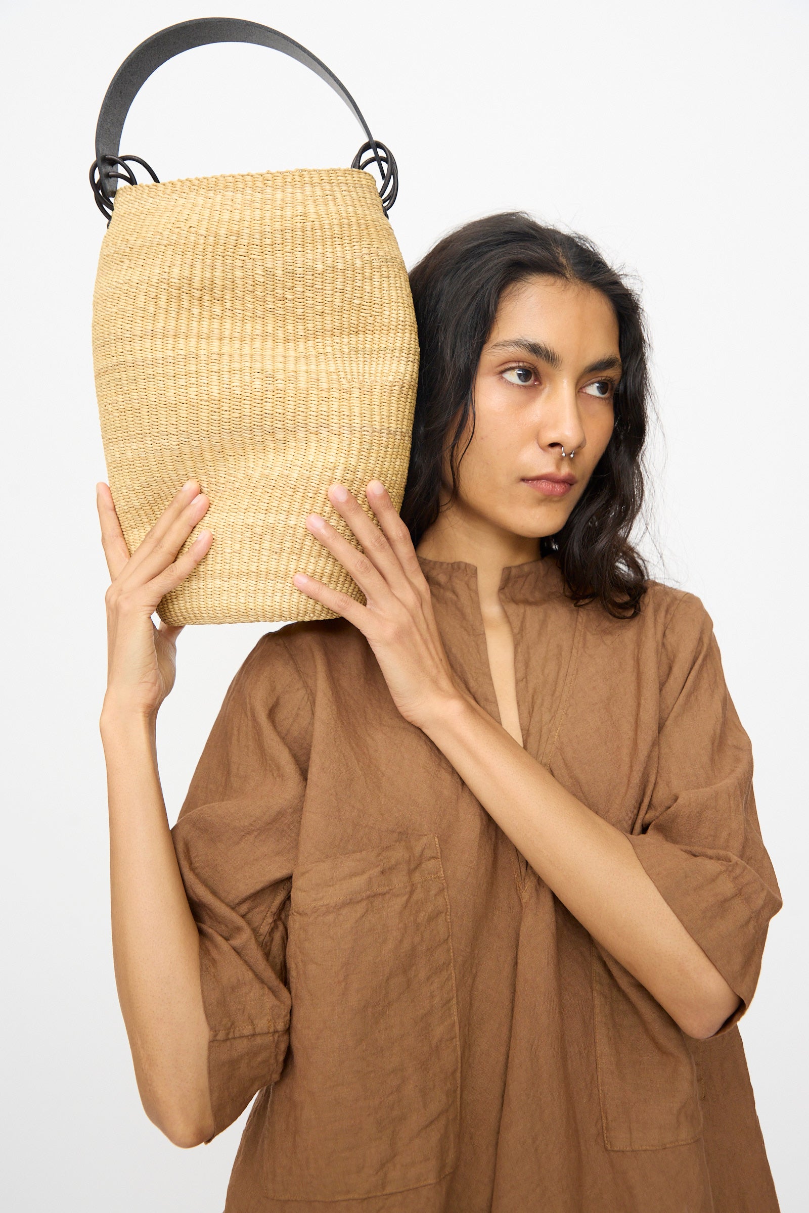 A person in a brown top holds the Inès Bressand Pot Froissé Bag in Natural, a handmade asymmetrical Ghanaian bucket bag with a black handle, next to their head against a plain white background.
