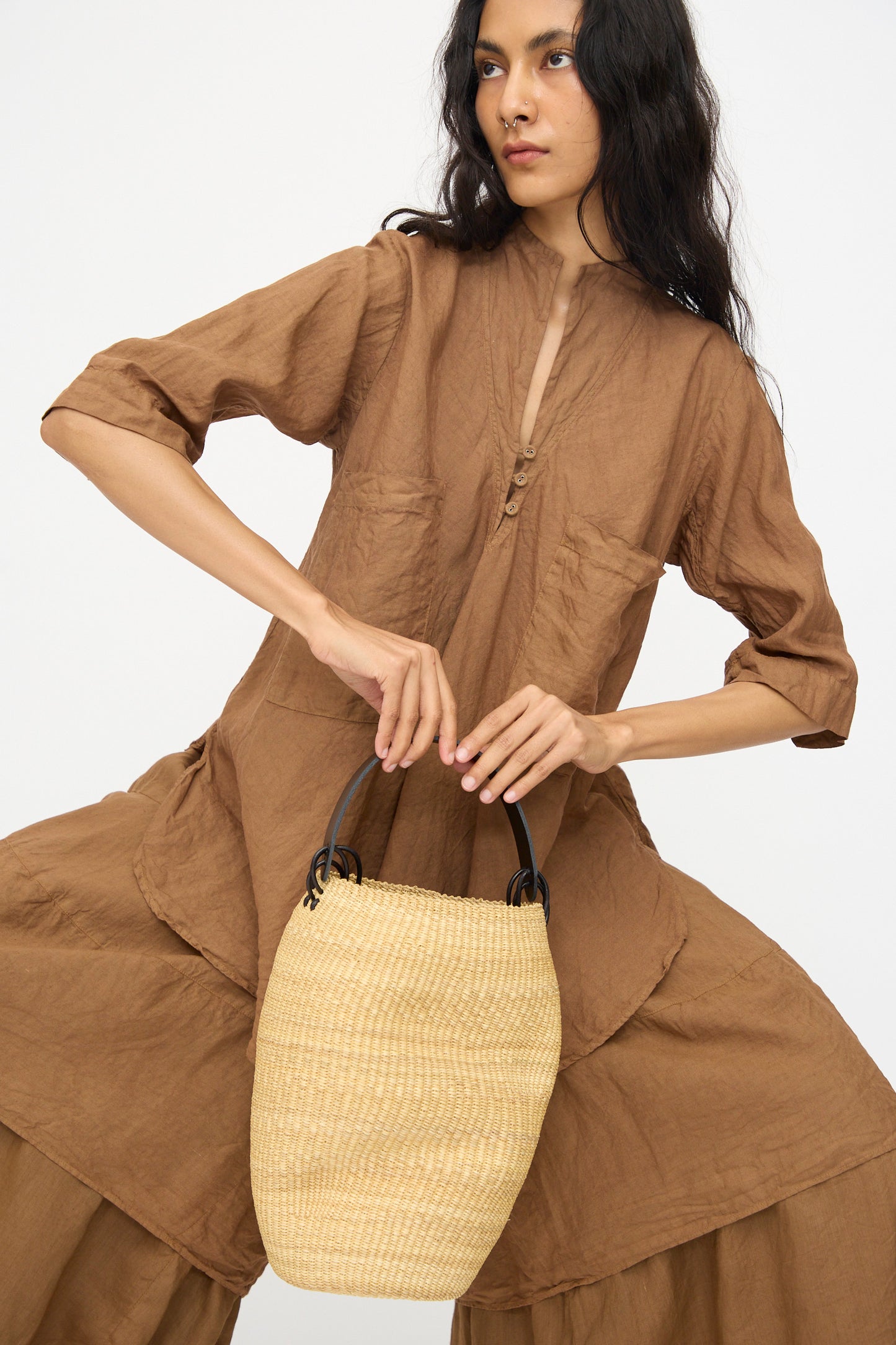 A woman in a loose brown outfit holds the Pot Froissé Bag in Natural by Inès Bressand, handmade in Ghana from elephant grass with black handles, against a plain white background.