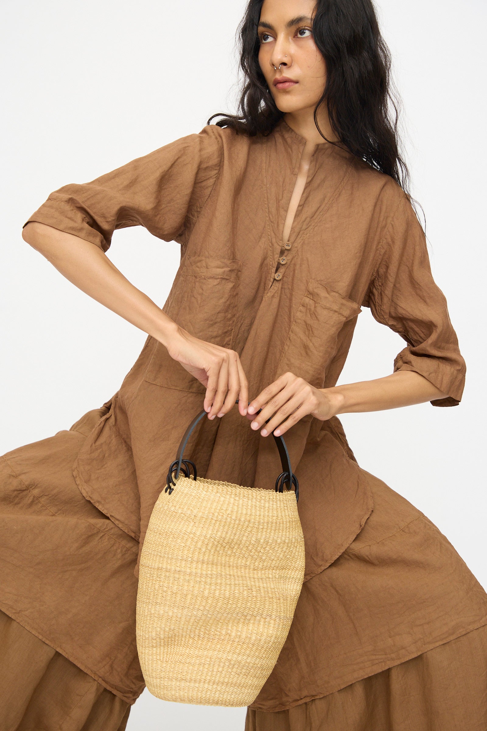 A woman in a loose brown outfit holds the Pot Froissé Bag in Natural by Inès Bressand, handmade in Ghana from elephant grass with black handles, against a plain white background.