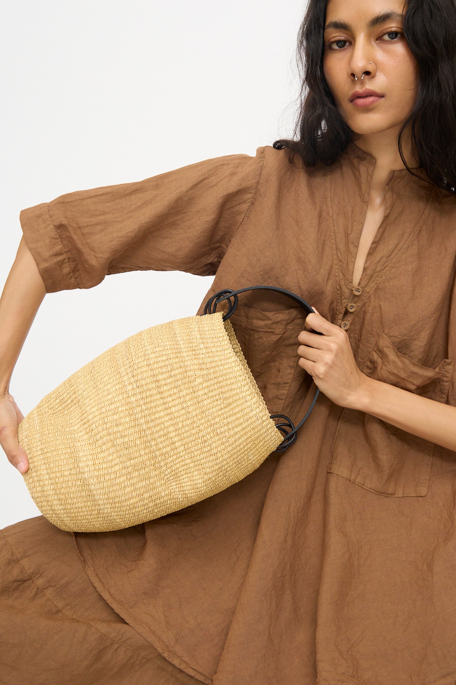 A person in a brown dress holds the Pot Froissé Bag in Natural by Inès Bressand, an asymmetrical woven straw bucket bag with black handles handmade in Ghana, against a plain light background.