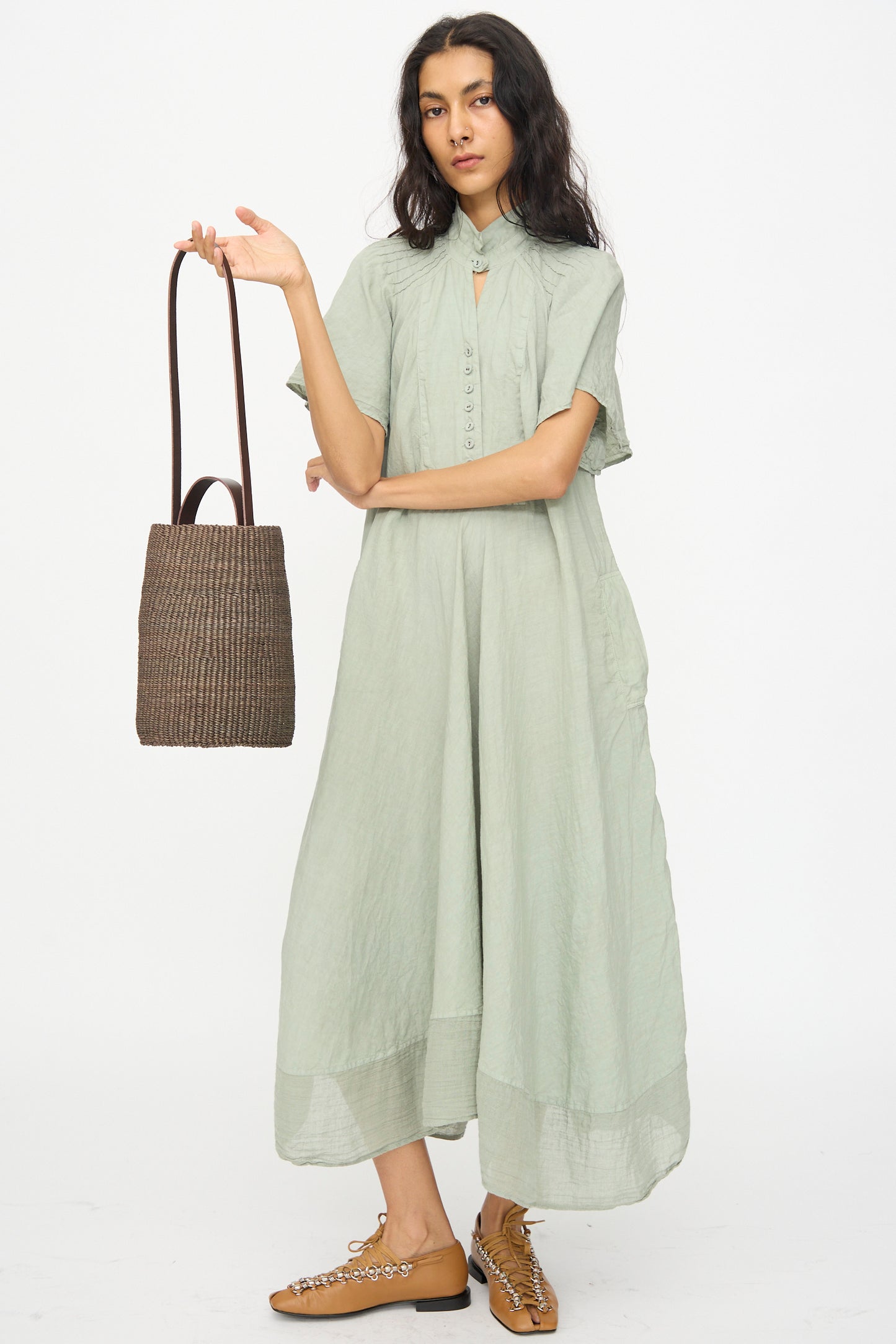 A person in a loose, light green short-sleeve dress holds the Inès Bressand Seau Bag in Brown and wears brown flat shoes, standing against a plain white background.