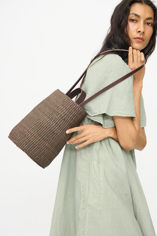 A woman in a light green dress carries the Inès Bressand Seau Bag in Brown, its strap resting on her shoulder against a plain white background.