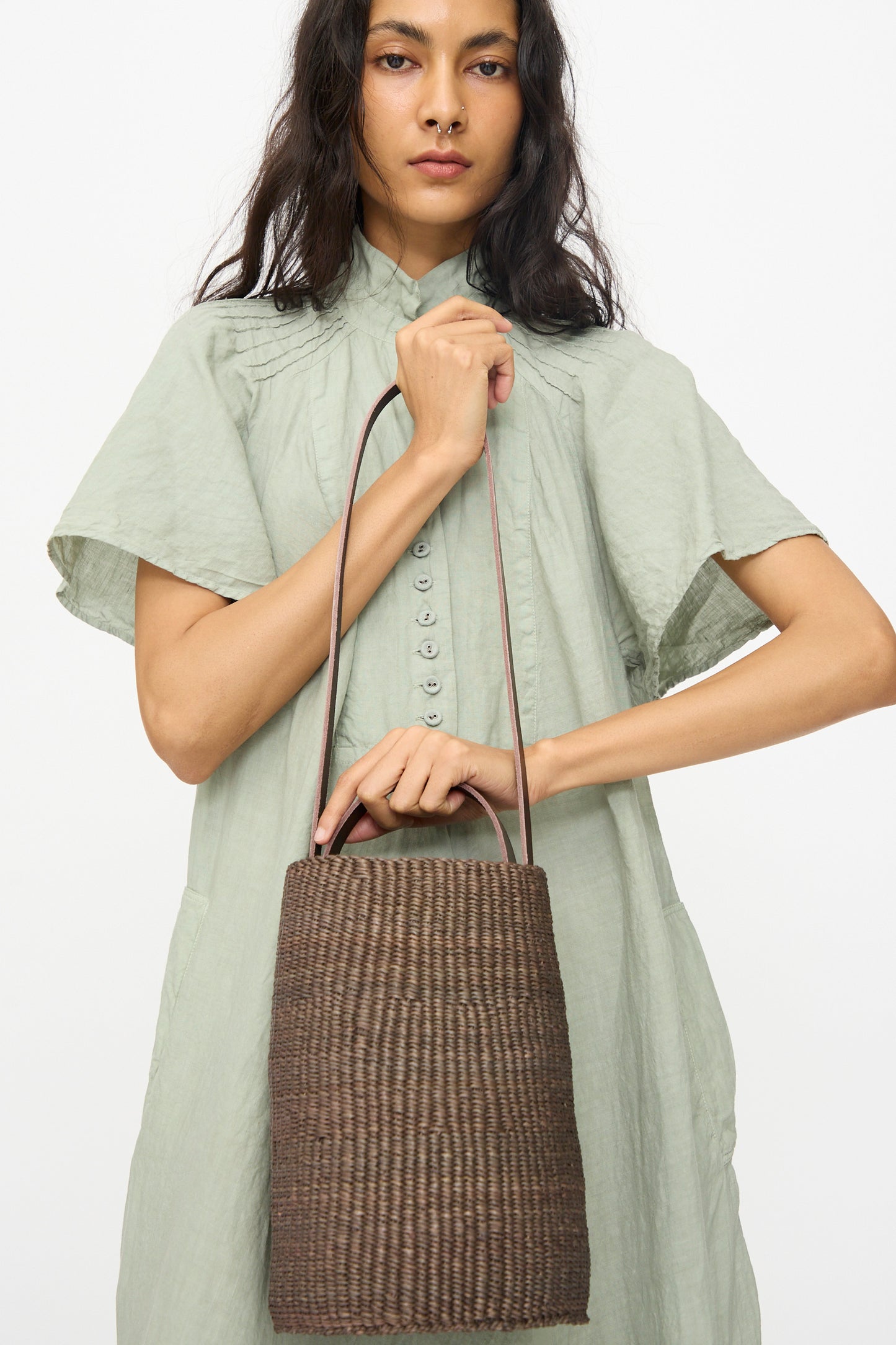 A woman holds the Inès Bressand Seau Bag in Brown, a handmade elephant grass bucket bag with two handles, while wearing a light green button-up dress against a plain white background.