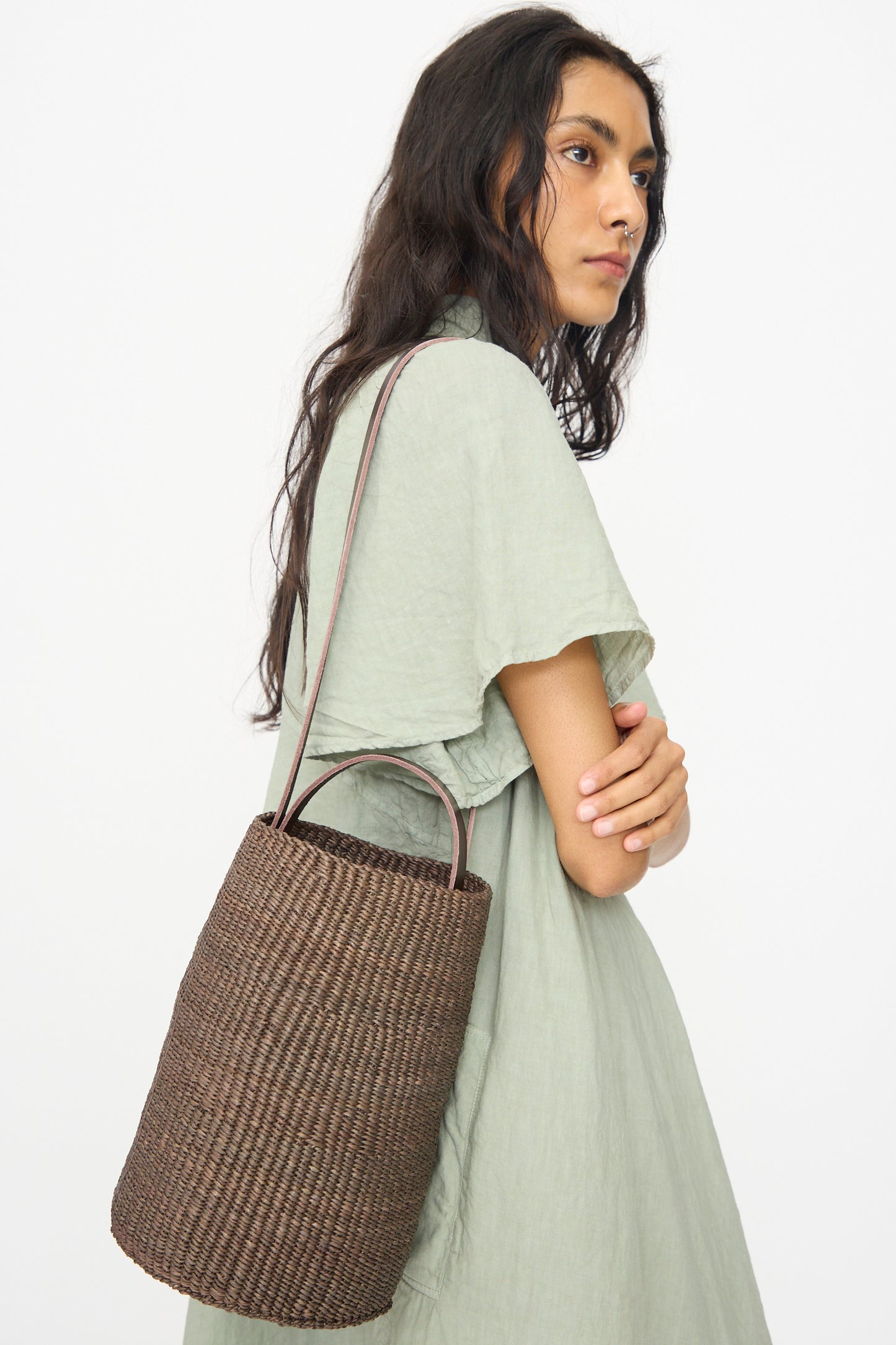 A woman in a light green dress stands sideways, holding the Inès Bressand Seau Bag in Brown with long handles against a plain white background.