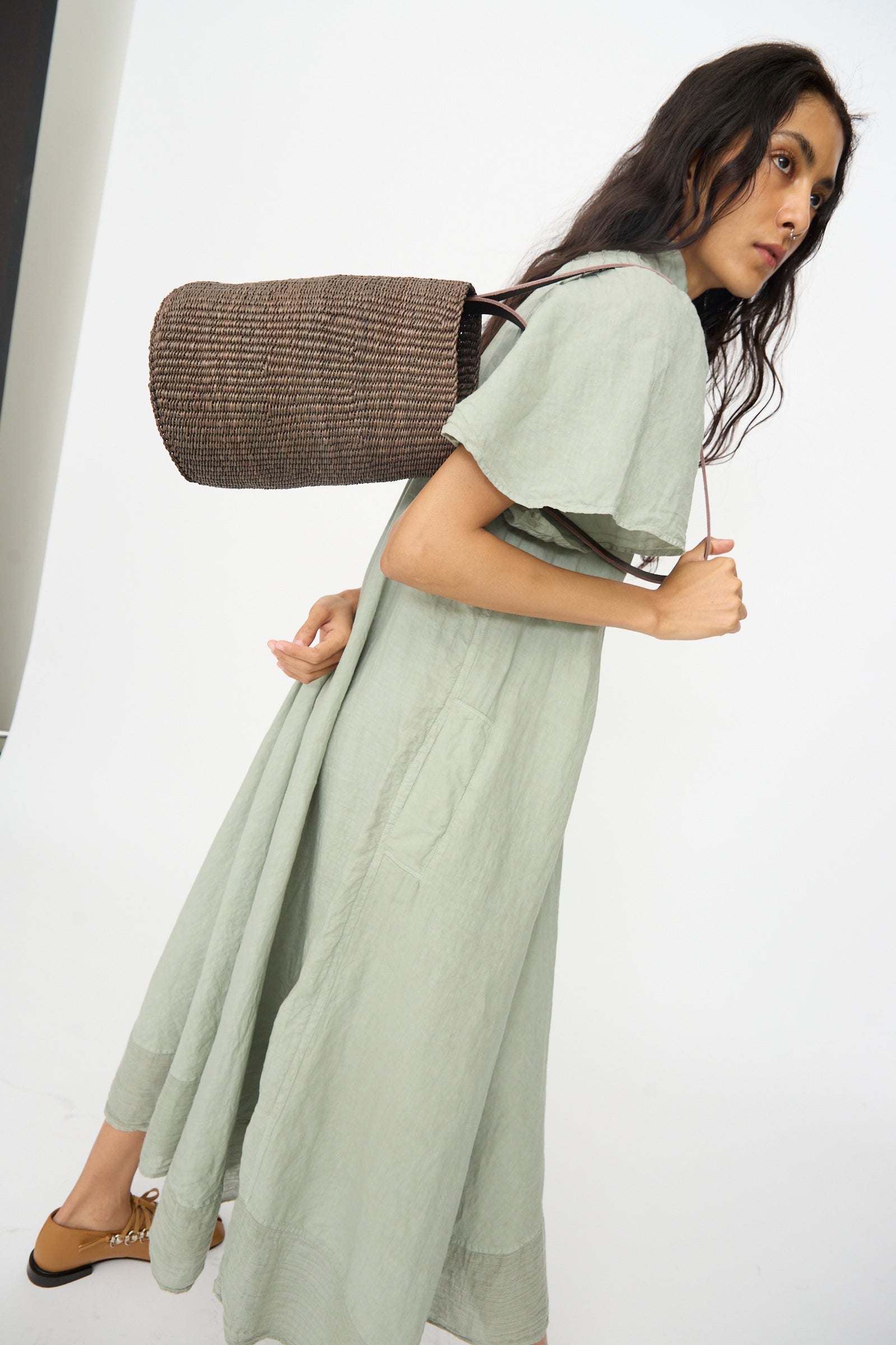A person in a light green dress stands sideways, carrying the Inès Bressand Seau Bag in Brown over one shoulder against a plain white background.