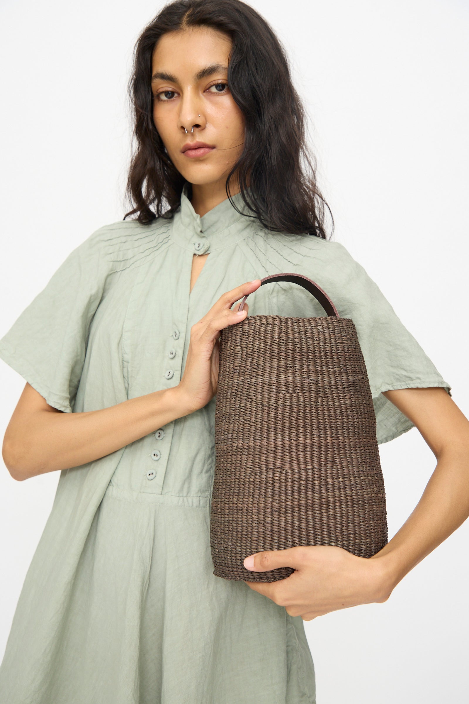 A woman in a light green buttoned dress holds the Seau Bag in Brown by Inès Bressand, a handmade bucket bag crafted from elephant grass, against a plain white background.