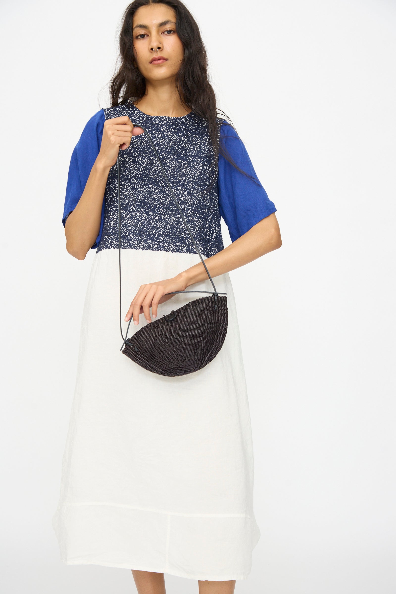 A woman in a blue and white dress holds the Inès Bressand Small Walnut Bag in Black, an oval crossbody with a long strap, against a plain white background.