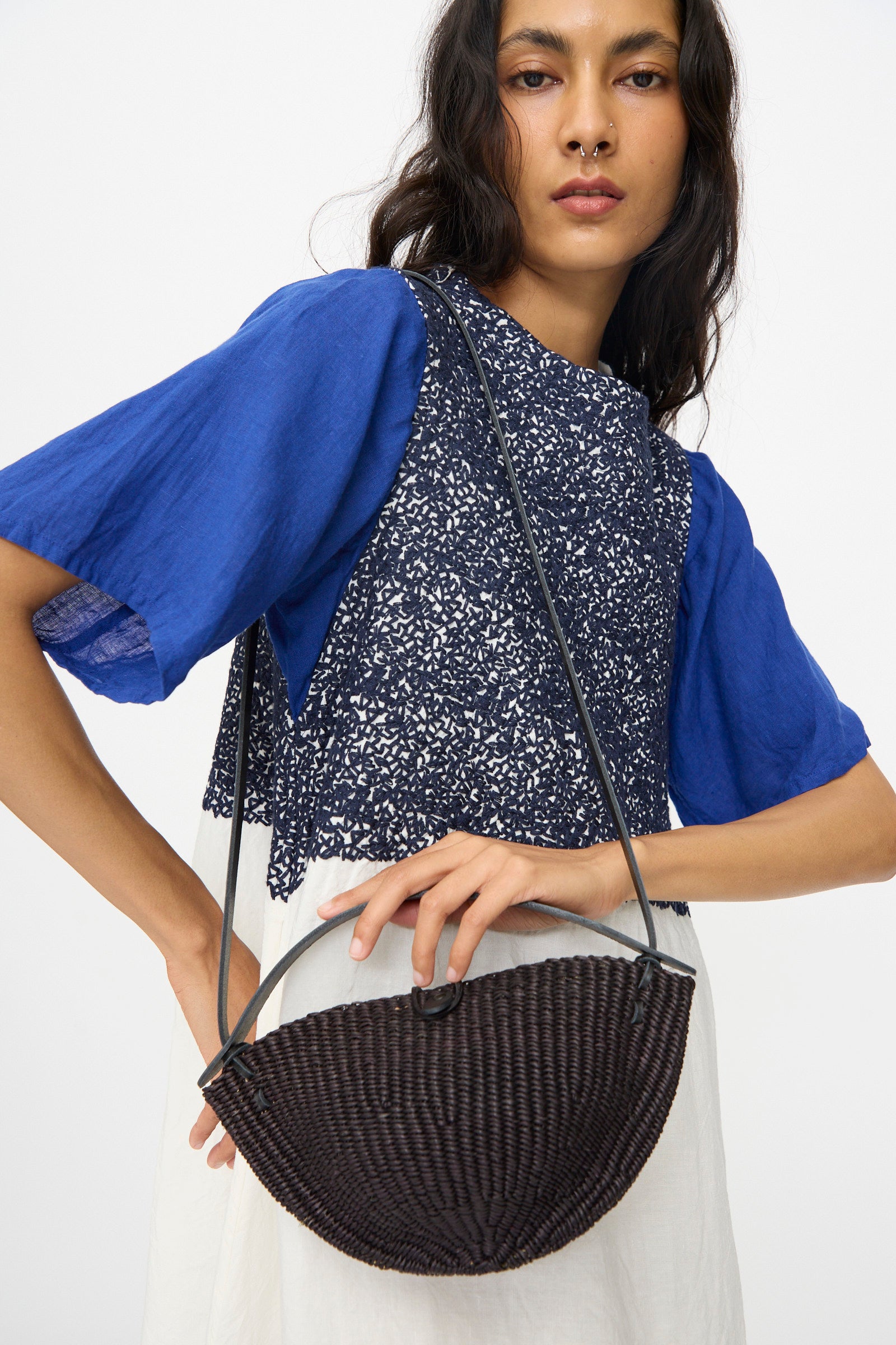 A woman with one hand on her hip carries the Small Walnut Bag in Black by Inès Bressand, an oval crossbody accessory skillfully handmade in Ghana from elephant grass.