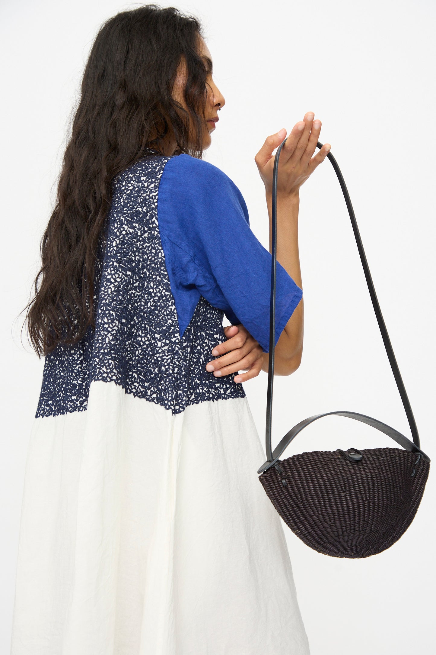 A person with long dark hair wears a blue and white patterned dress and holds the Small Walnut Bag in Black by Inès Bressand against a plain white background.