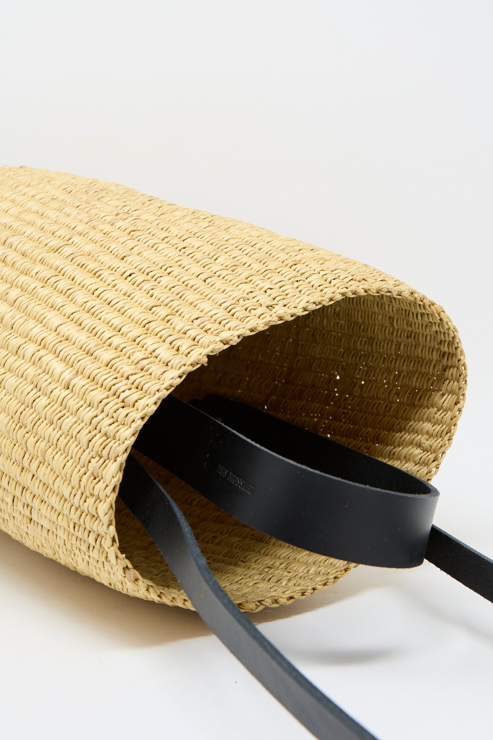 The Seau Bag in Natural with Black by Inès Bressand, handcrafted from elephant grass in Ghana, features two extending black leather straps as it rests on its side.