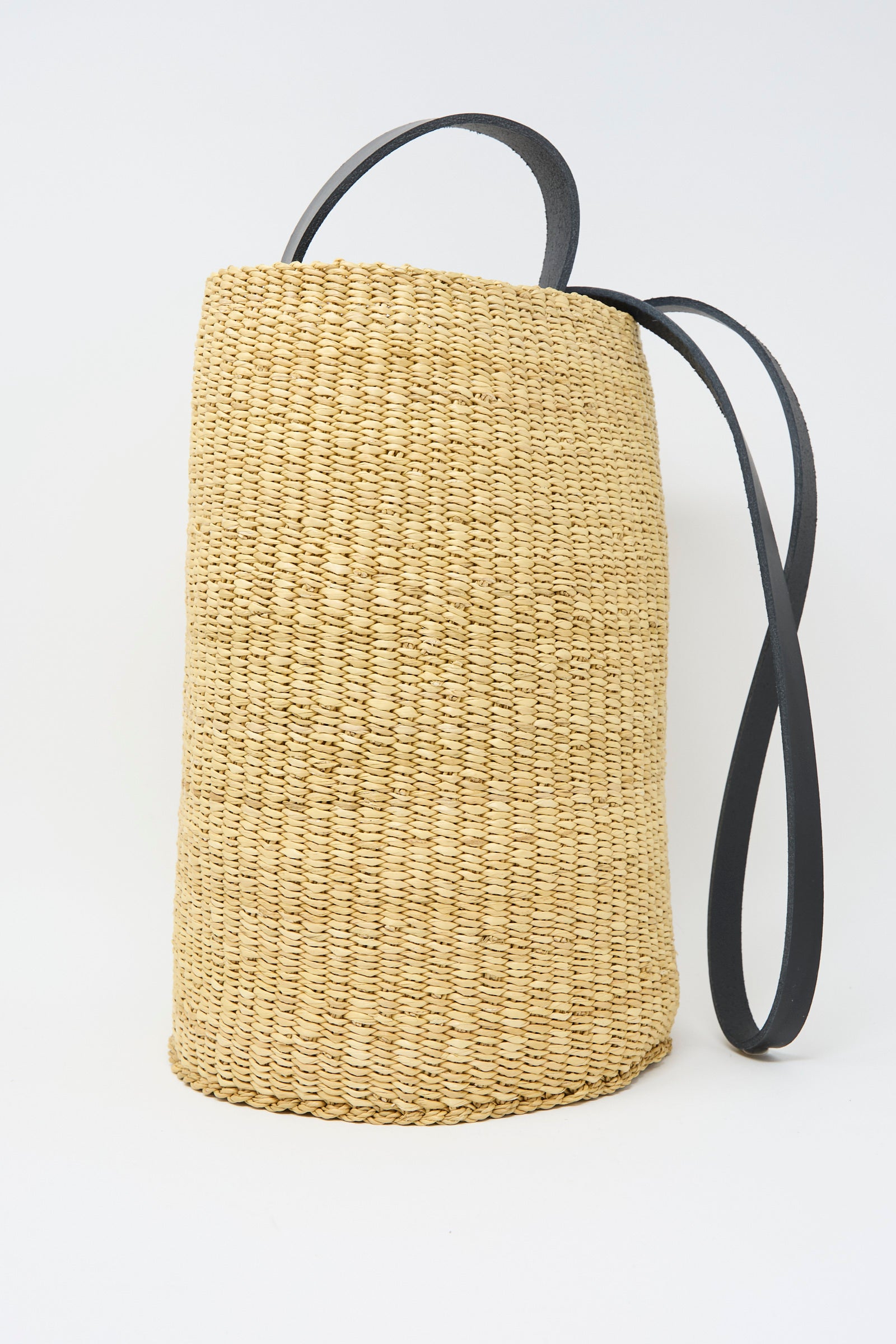 The Seau Bag in Natural with Black by Inès Bressand, made from handwoven elephant grass with a black leather shoulder strap, sits against a plain white backdrop.