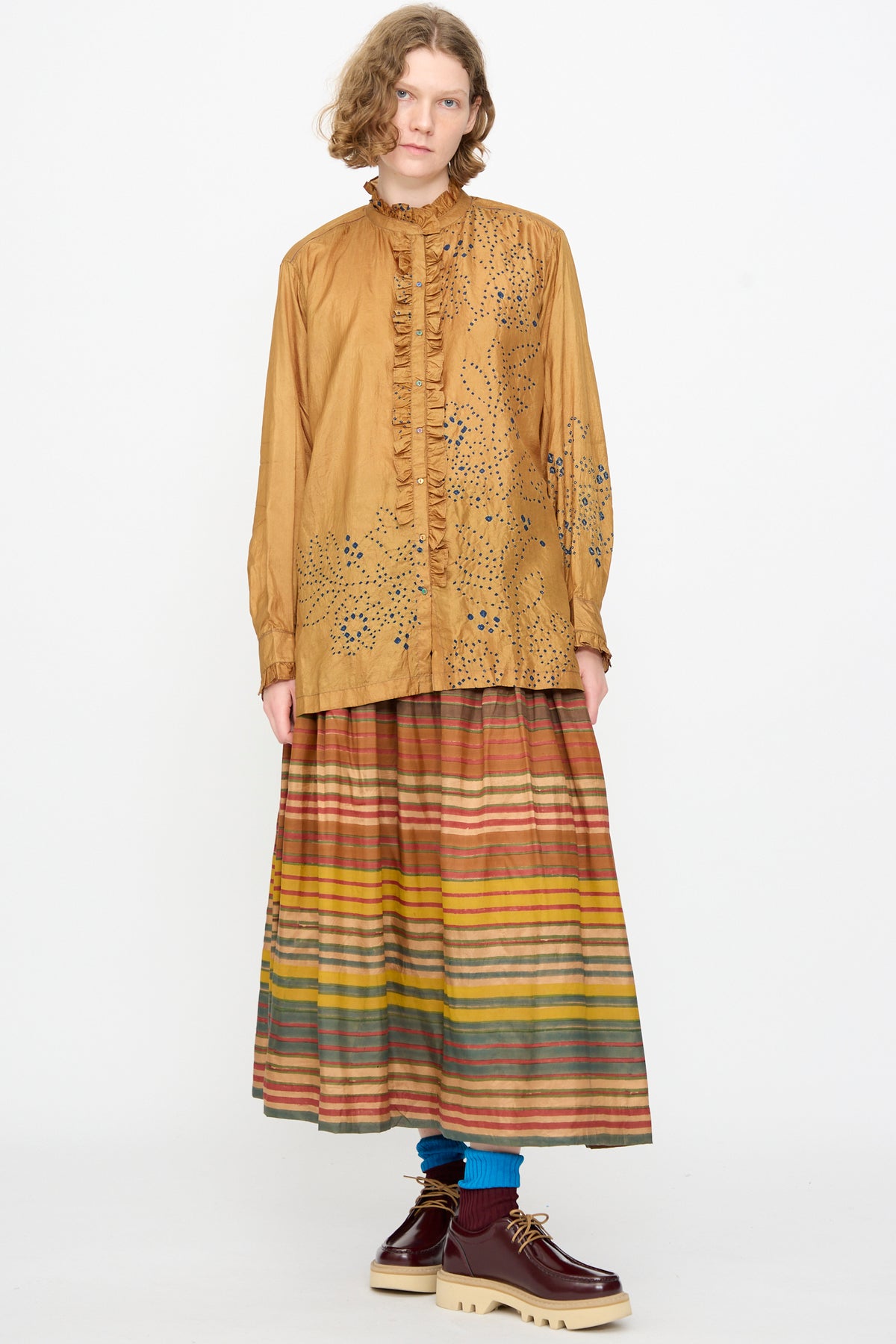 Wearing the Bandhani Silk Henri Shirt in Ochre by Injiri, paired with a pleated striped skirt in earth tones, blue socks, and burgundy platform shoes, the person stands against a plain white background.