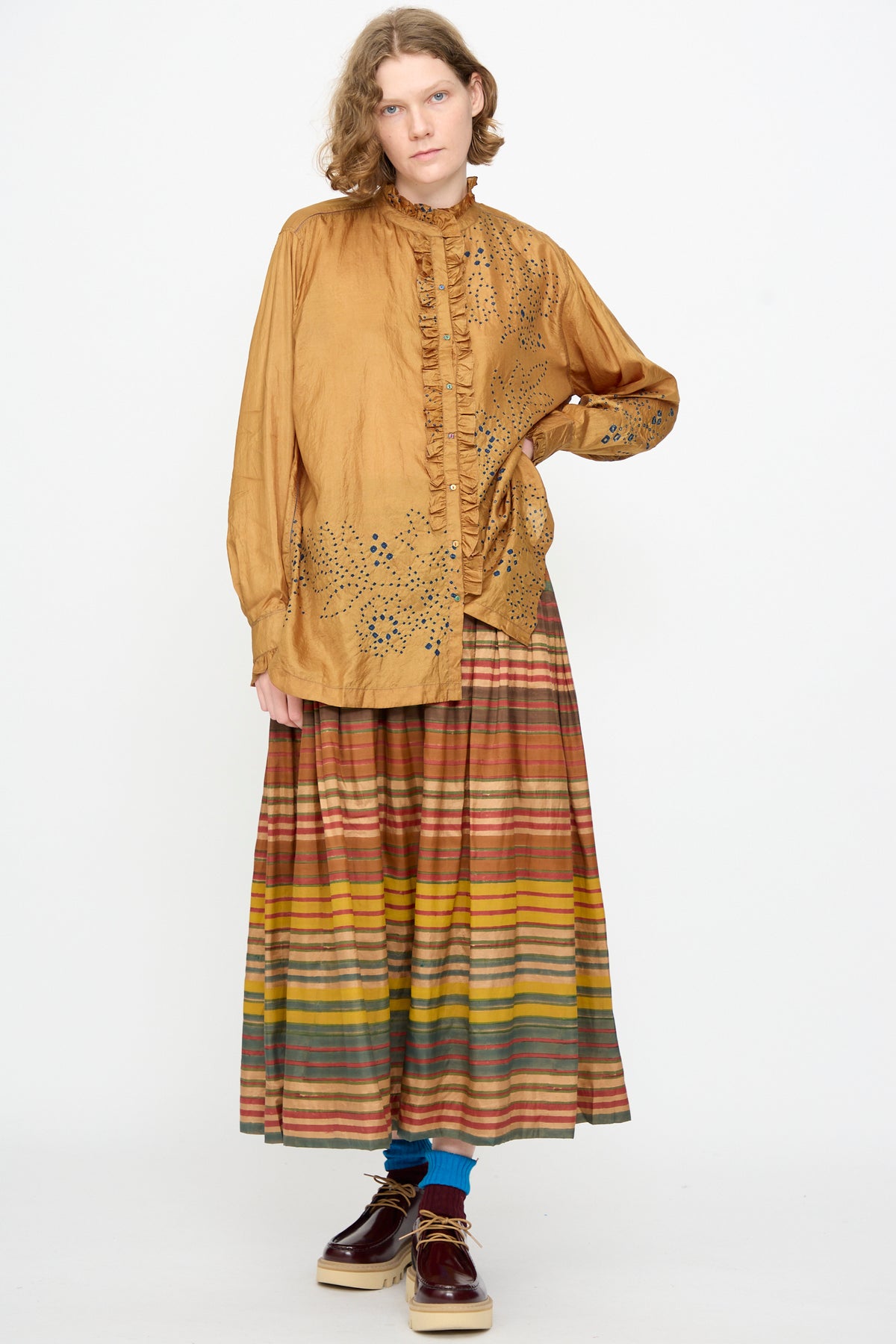 Wearing the Injiri Bandhani Silk Henri Shirt in Ochre, featuring a loose silhouette and a dotted pattern, paired with a long, striped skirt in warm hues, blue socks, and brown platform shoes, the person stands against a plain white background.