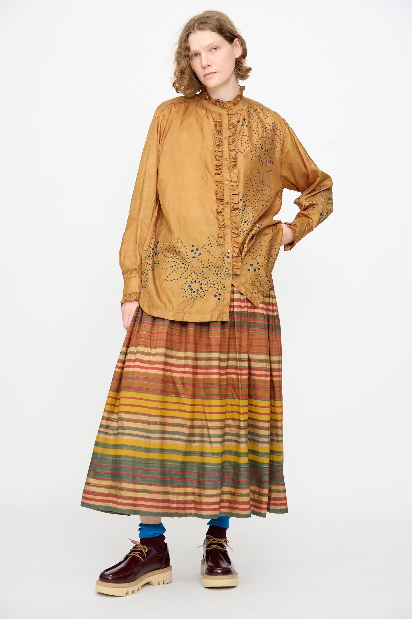 Wearing the Injiri Bandhani Silk Henri Shirt in Ochre, a loose, handcrafted silk top adorned with decorative dots, paired with a long, multicolored striped skirt, blue socks, and brown platform shoes—standing against a plain white background.