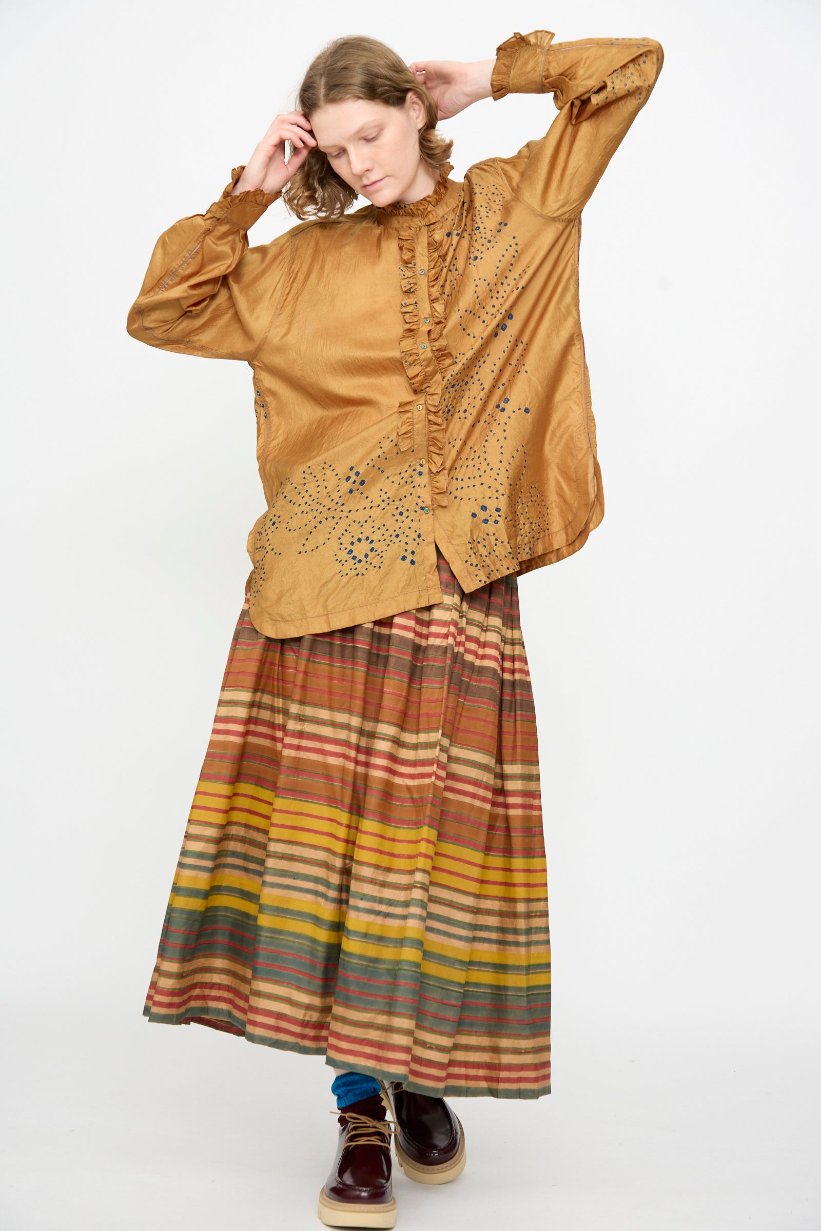 A person wearing a loose, ochre Bandhani Silk Henri Shirt by Injiri with ruffled accents and laser-cut details, paired with a long, multicolored striped skirt, standing against a plain white background.