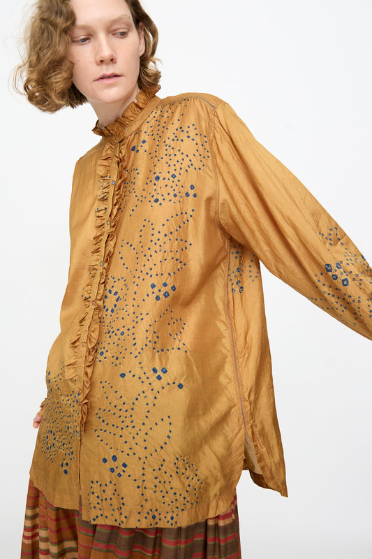A person wearing the Injiri Bandhani Silk Henri Shirt in Ochre—a loose-fitting shirt with ruffle trim and blue dotted motifs—paired with a striped skirt.