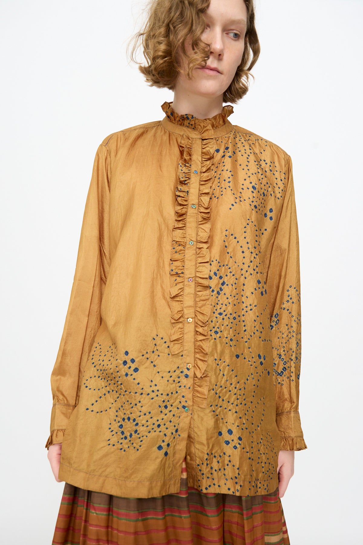 A person wearing the Injiri Bandhani Silk Henri Shirt in Ochre—a loose, handcrafted silk shirt featuring ruffles and blue dotted patterns—paired with a striped skirt, stands against a plain background.