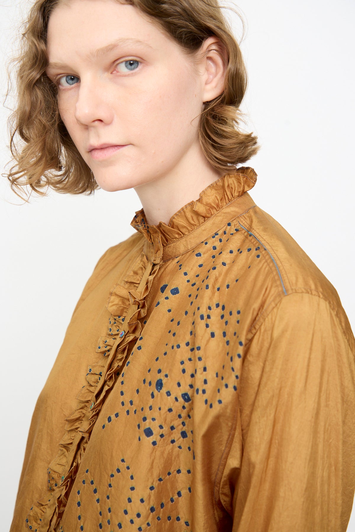 Person with short, wavy light brown hair wearing the Injiri Bandhani Silk Henri Shirt in Ochre—an artisanal piece crafted from silk featuring intricate blue dotted patterns and delicate ruffles—looking toward the camera against a plain background.