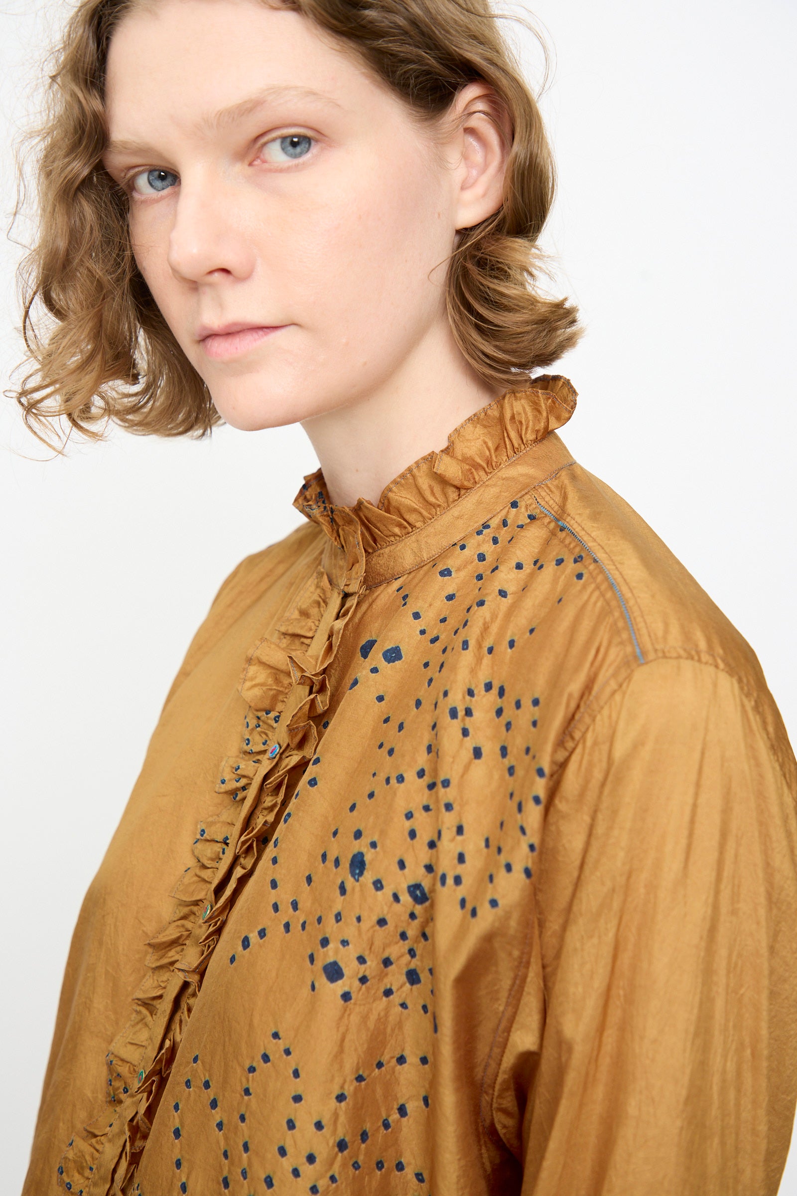 Person with short, wavy light brown hair wearing the Injiri Bandhani Silk Henri Shirt in Ochre—an artisanal piece crafted from silk featuring intricate blue dotted patterns and delicate ruffles—looking toward the camera against a plain background.