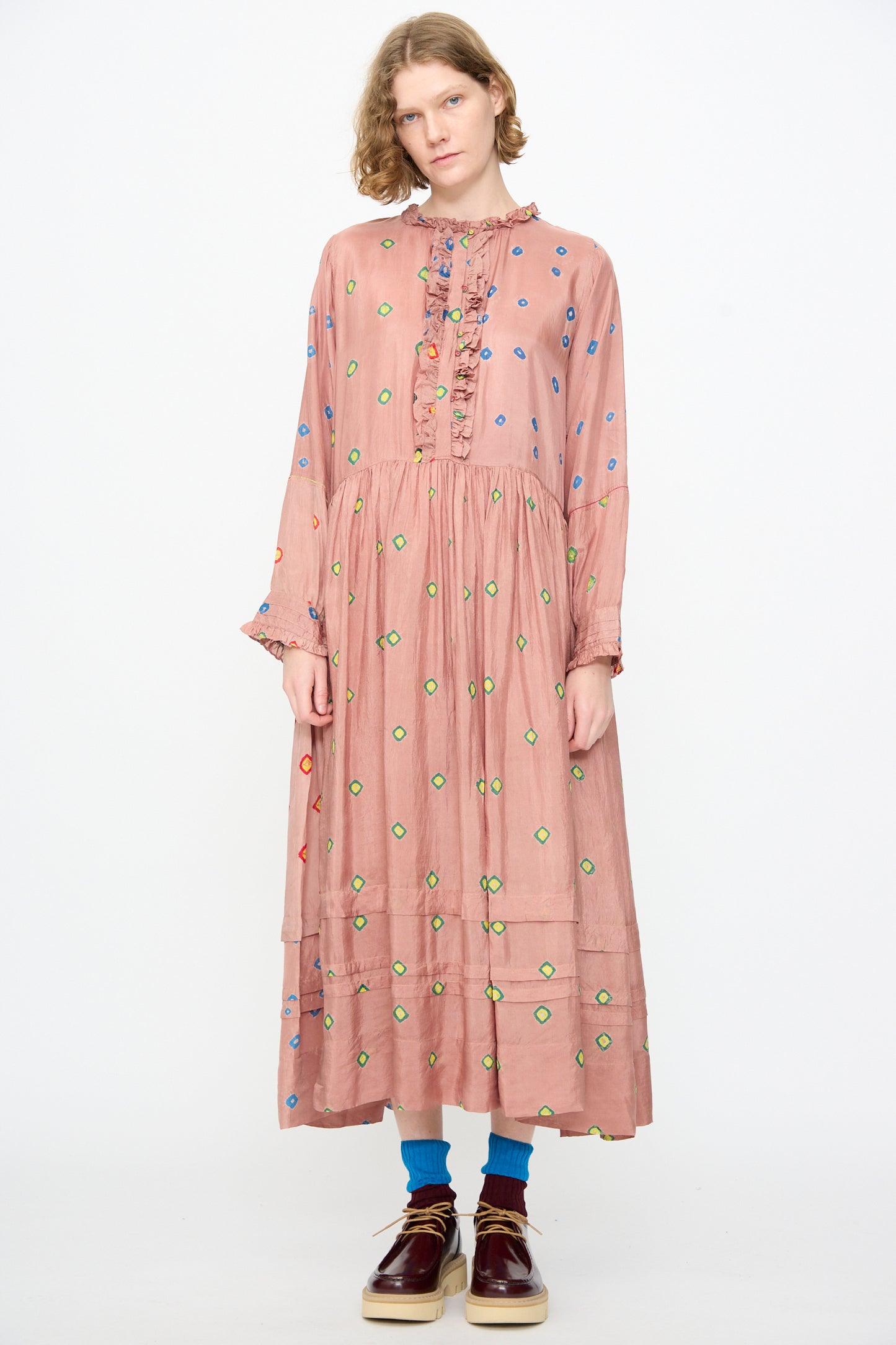 A person wearing the Injiri Bandhani Silk Natalie Dress in Mauve—a long, handcrafted maxi dress with ruffle details and colorful polka dots—paired with blue socks and brown shoes, standing against a plain white background.