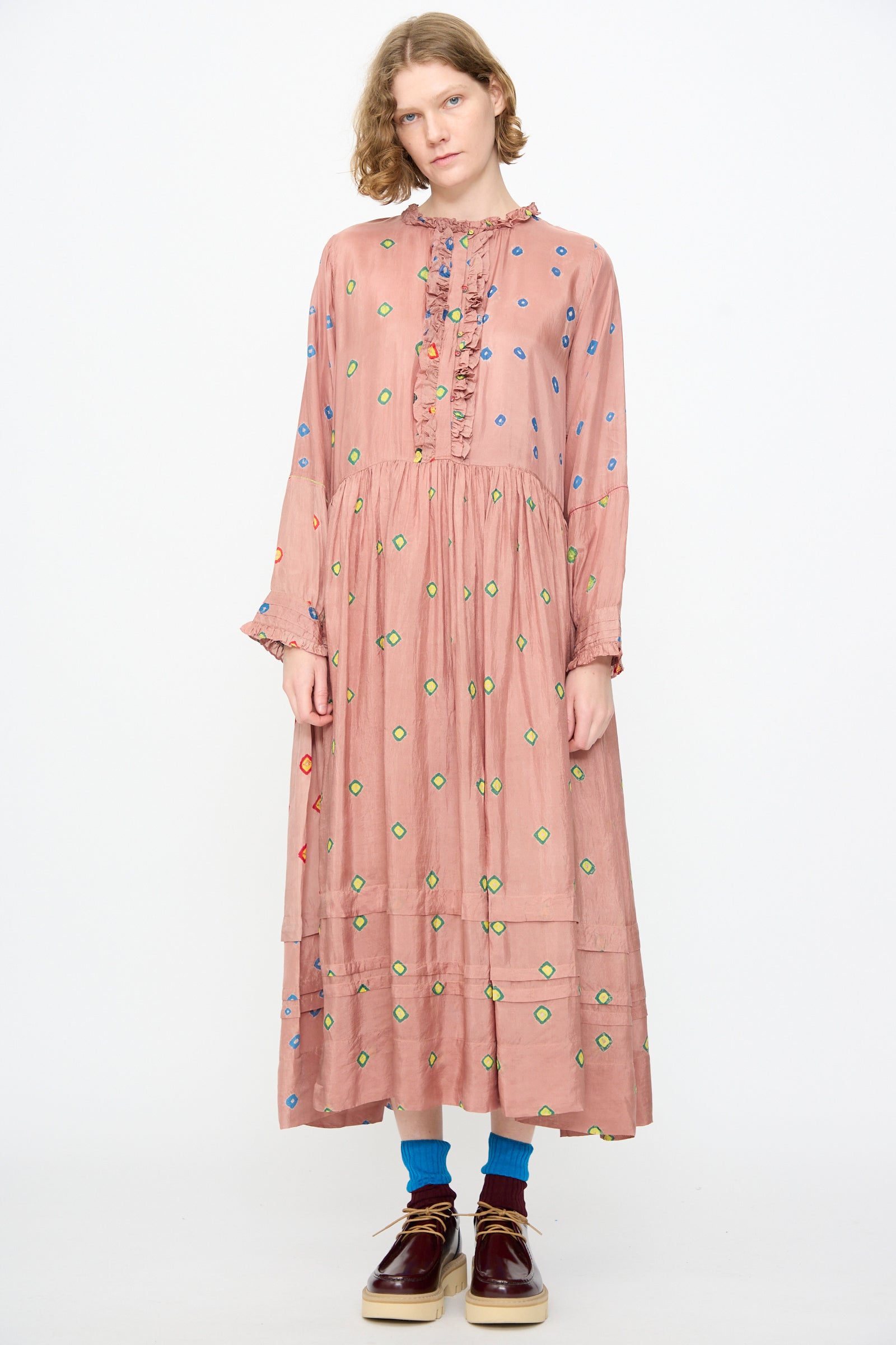 A person wearing the Injiri Bandhani Silk Natalie Dress in Mauve—a long, handcrafted maxi dress with ruffle details and colorful polka dots—paired with blue socks and brown shoes, standing against a plain white background.
