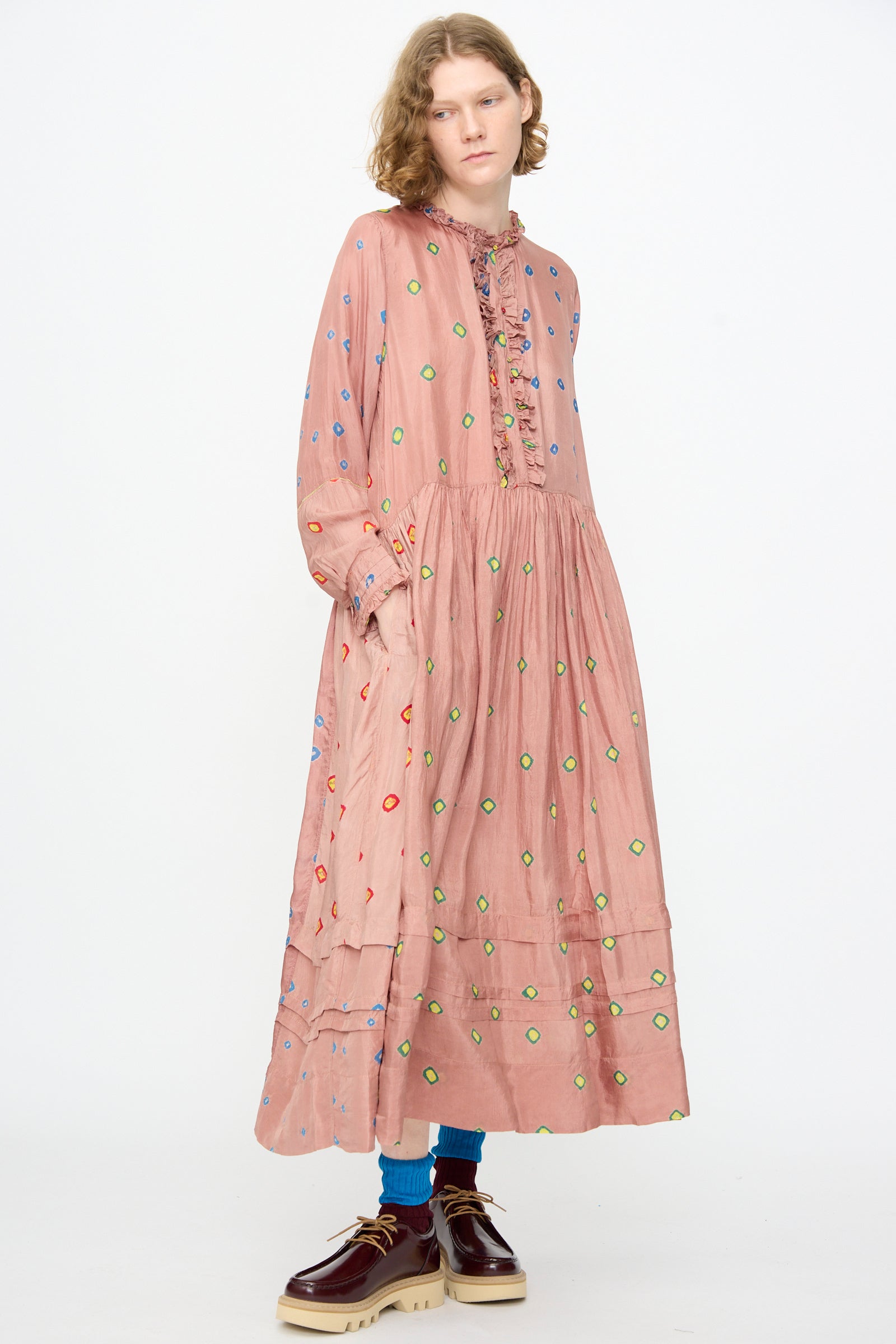 A person is wearing the Injiri Bandhani Silk Natalie Dress in Mauve—a loose, long pink tie-dyed silk dress with colorful circular patterns—paired with blue socks and brown platform shoes, standing against a plain white background.