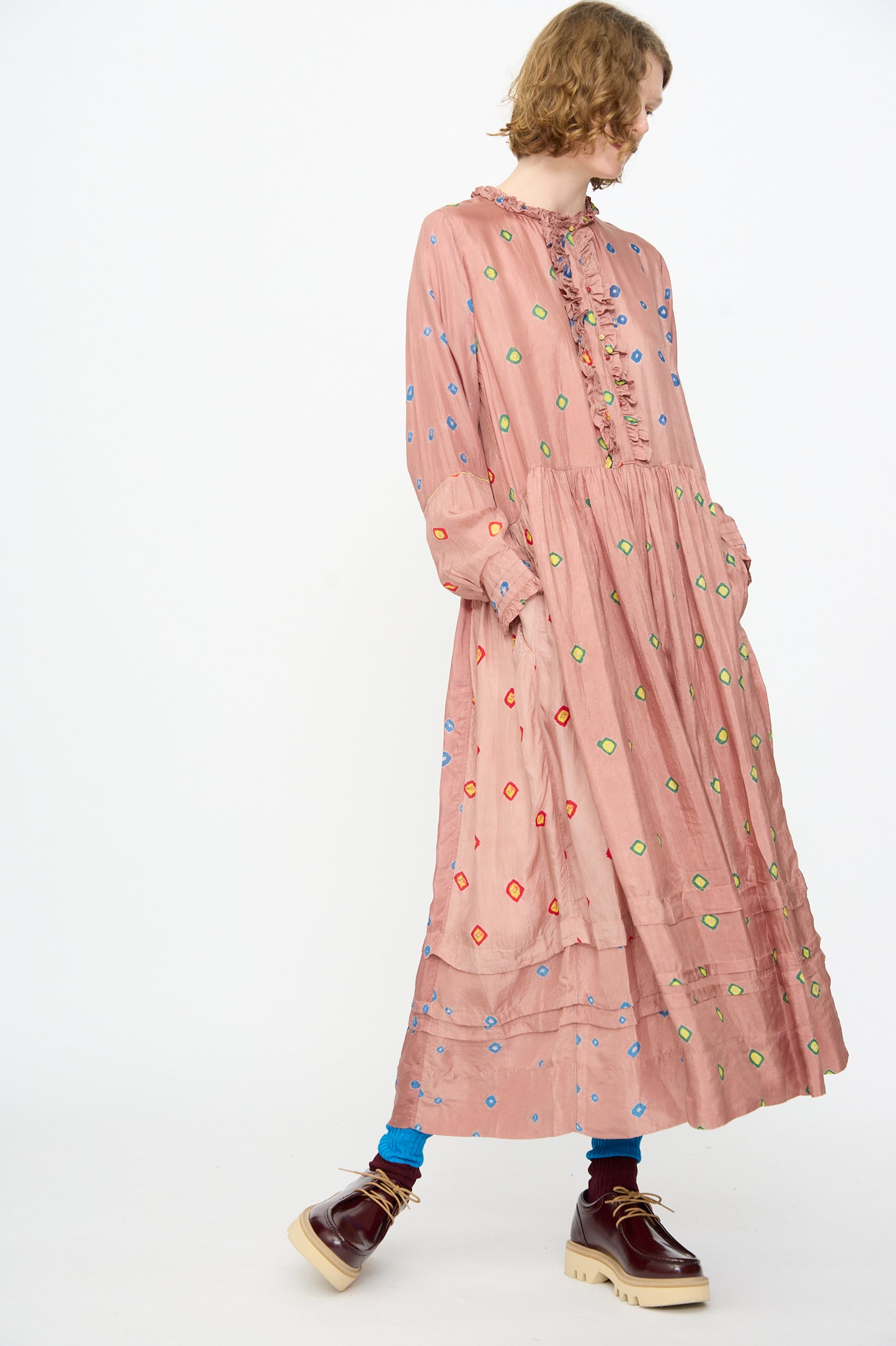 A person wearing the Bandhani Silk Natalie Dress in Mauve by Injiri—a long, loose-fitting handcrafted maxi dress in mauve adorned with colorful embroidered dots—paired with blue socks and burgundy platform shoes, standing against a white background.