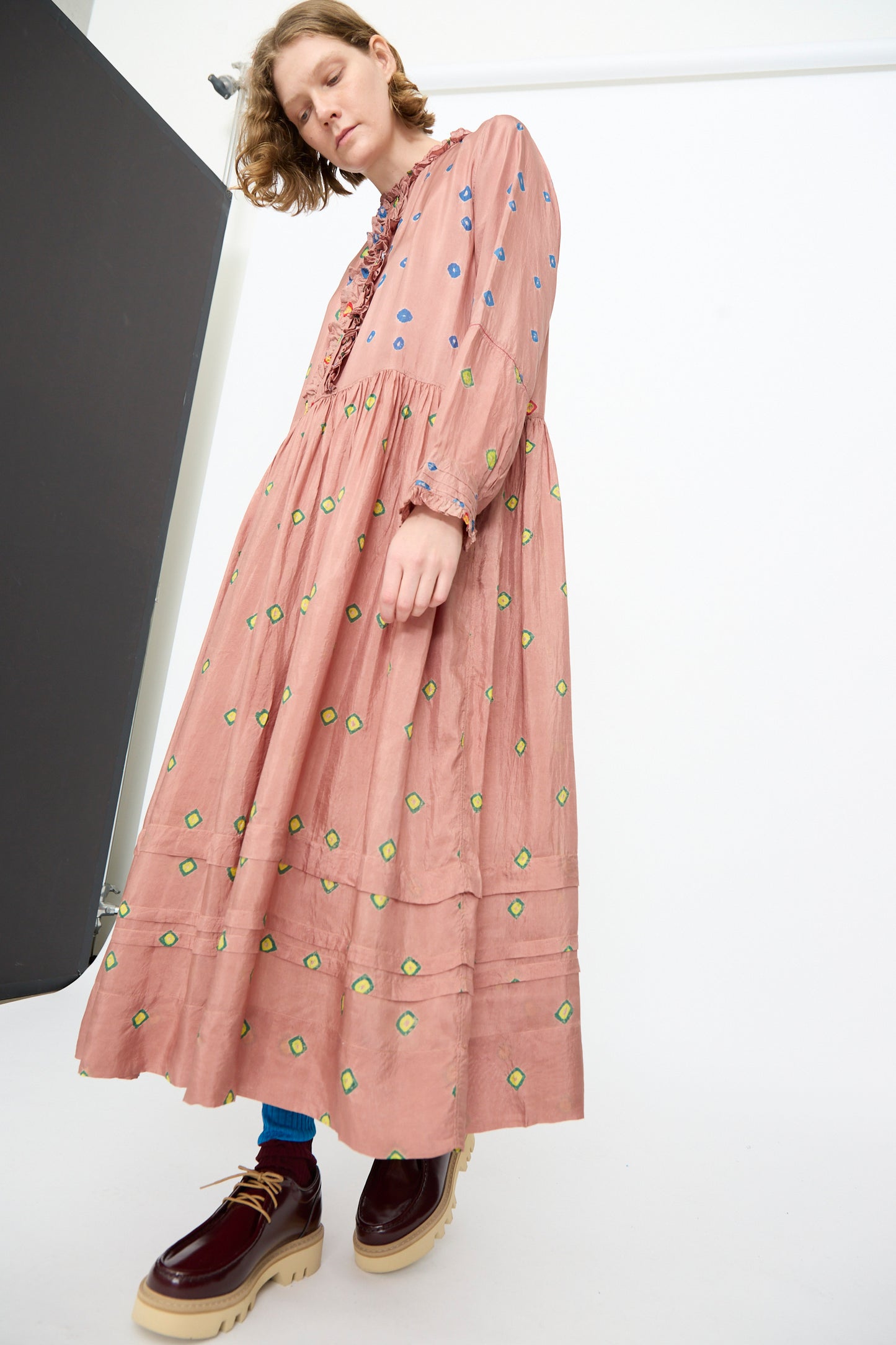 A person stands in a minimalist studio setting wearing the Injiri Bandhani Silk Natalie Dress in Mauve, featuring green and blue patterns, paired with maroon platform shoes.