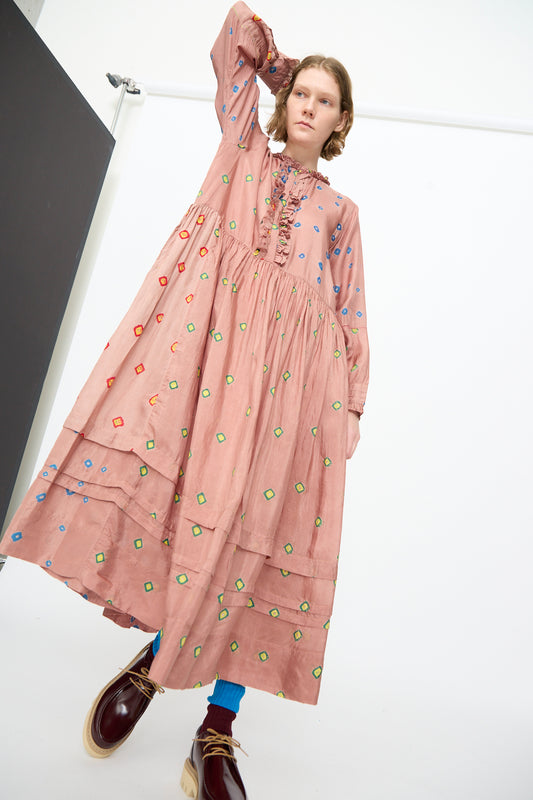 A person wearing the Injiri Bandhani Silk Natalie Dress in Mauve, featuring colorful geometric patterns, paired with blue tights and brown platform shoes, stands in a white room looking at the camera.