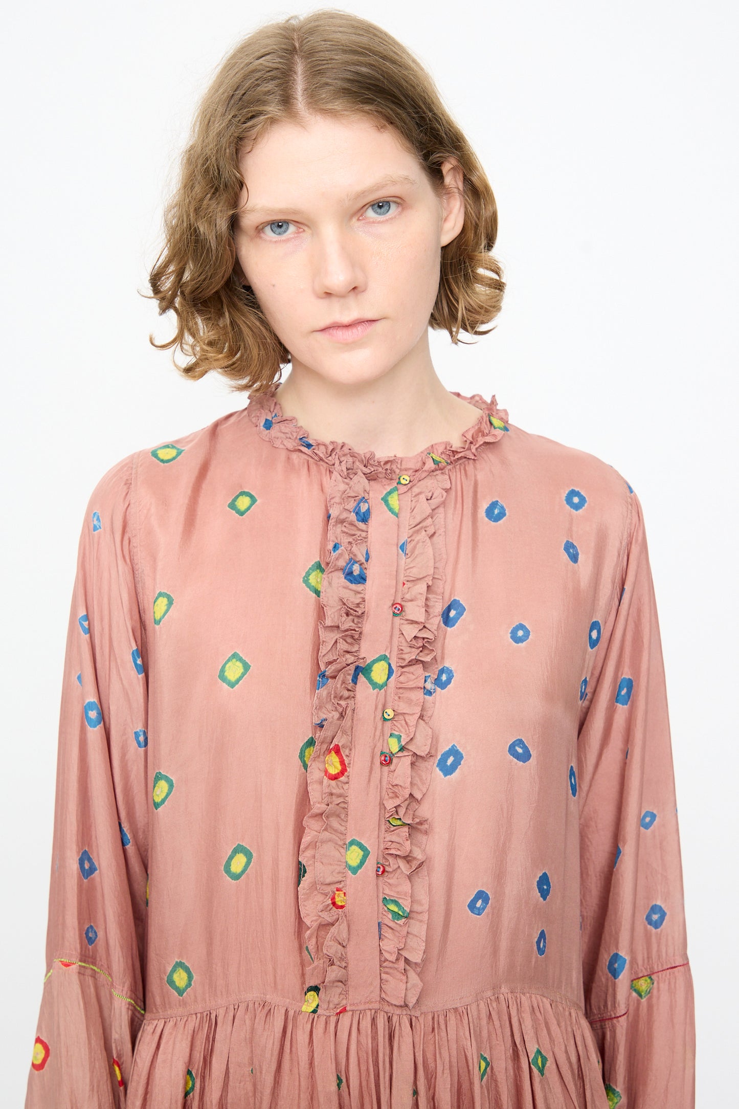 A person with short, wavy hair wears the Injiri Bandhani Silk Natalie Dress in Mauve—a handcrafted maxi dress adorned with colorful geometric patterns and ruffled detailing—standing against a plain white background.