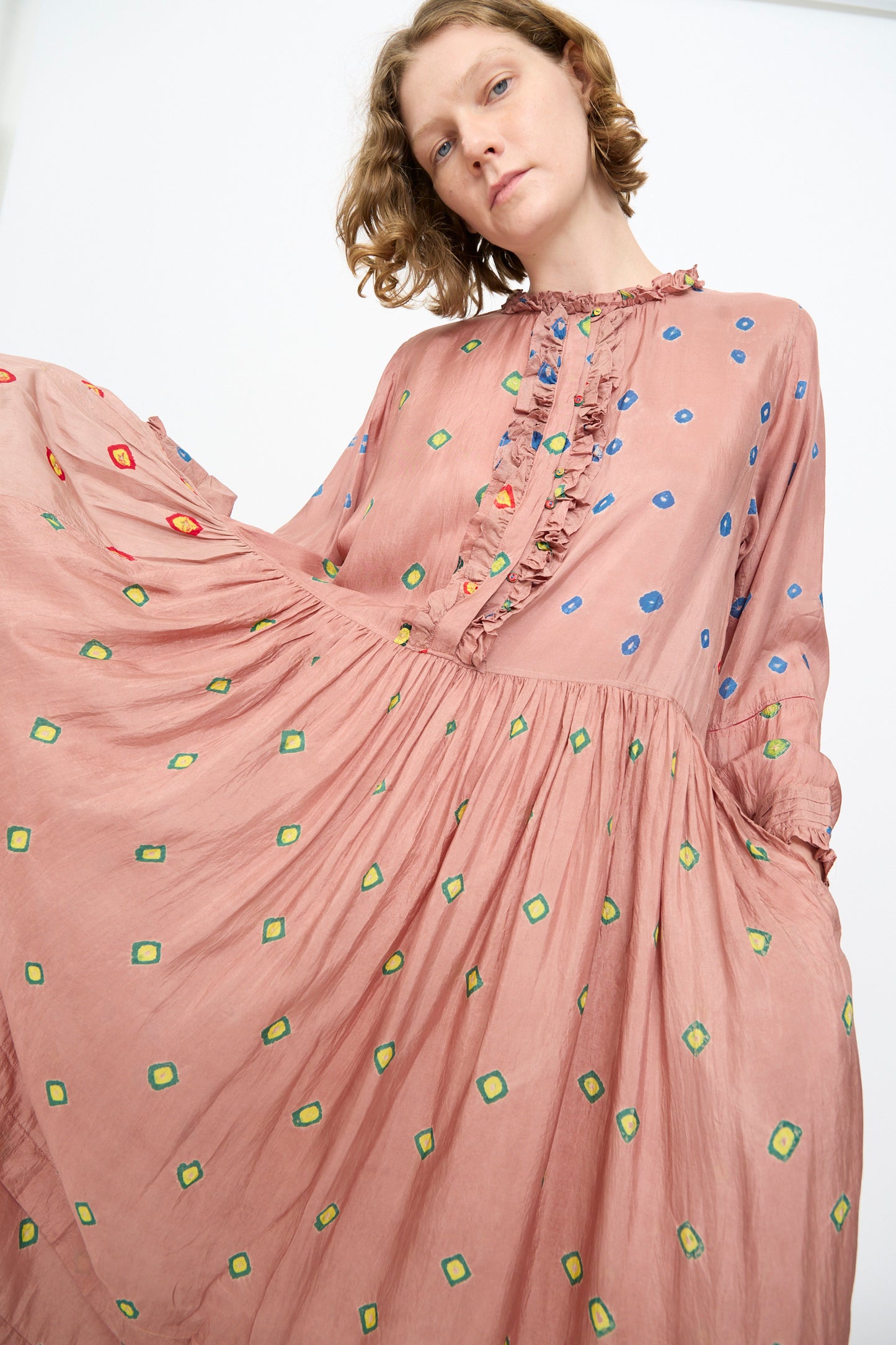 A person wearing the Injiri Bandhani Silk Natalie Dress in Mauve—a loose, handcrafted maxi dress featuring ruffles and colorful geometric patterns—holds out one side of the skirt against a plain background.