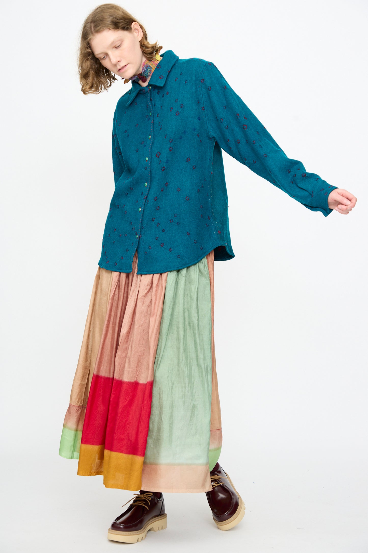 A person wearing the Injiri Bandhani Wool Shirt in Blue, handcrafted in India, paired with a long multicolored skirt featuring tiers of green, pink, and red, and brown platform shoes, stands against a plain white background.