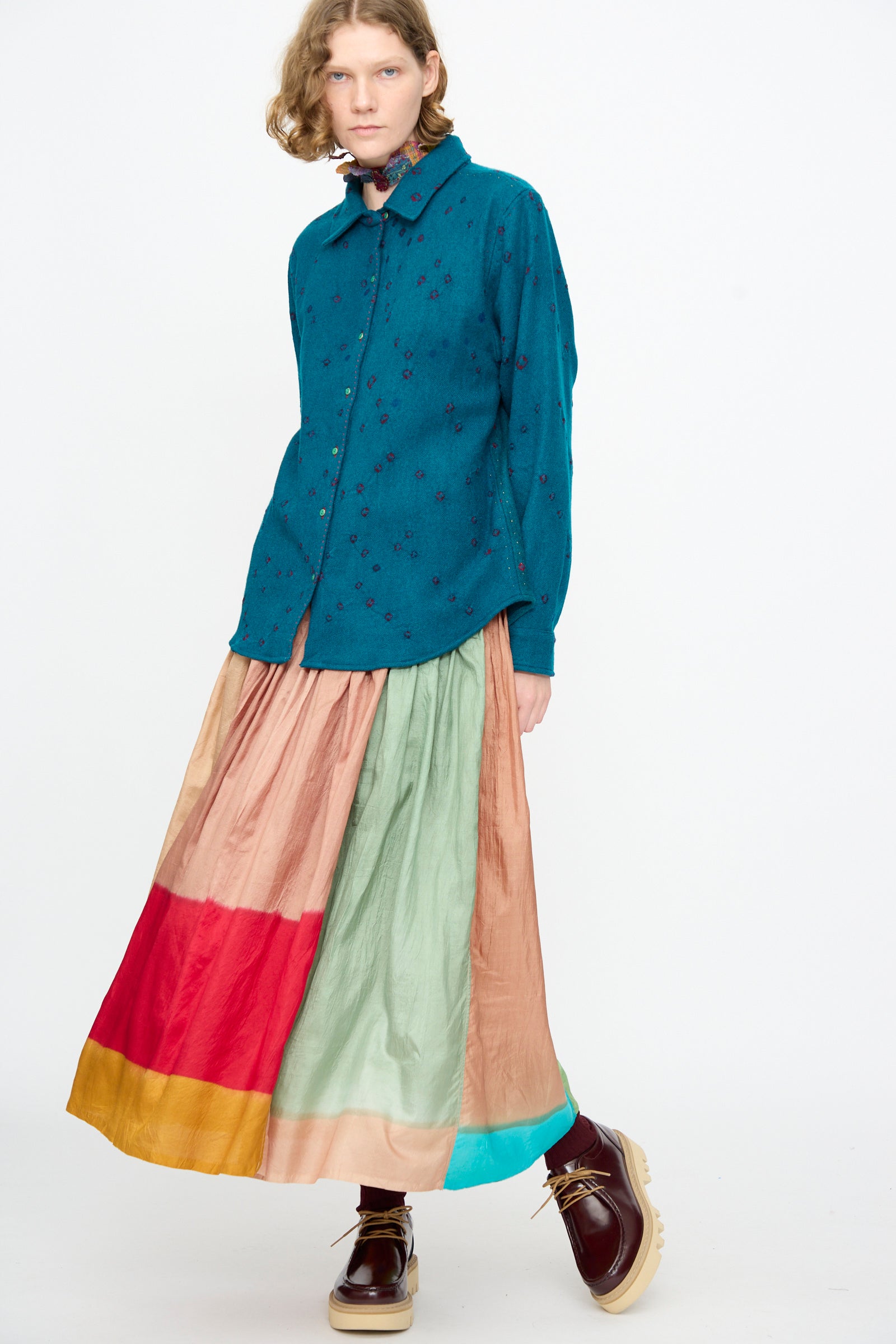 A person wearing the Injiri Bandhani Wool Shirt in Blue, handcrafted in India with a subtle pattern, paired with a long multicolored skirt and brown platform shoes, stands against a plain white background.