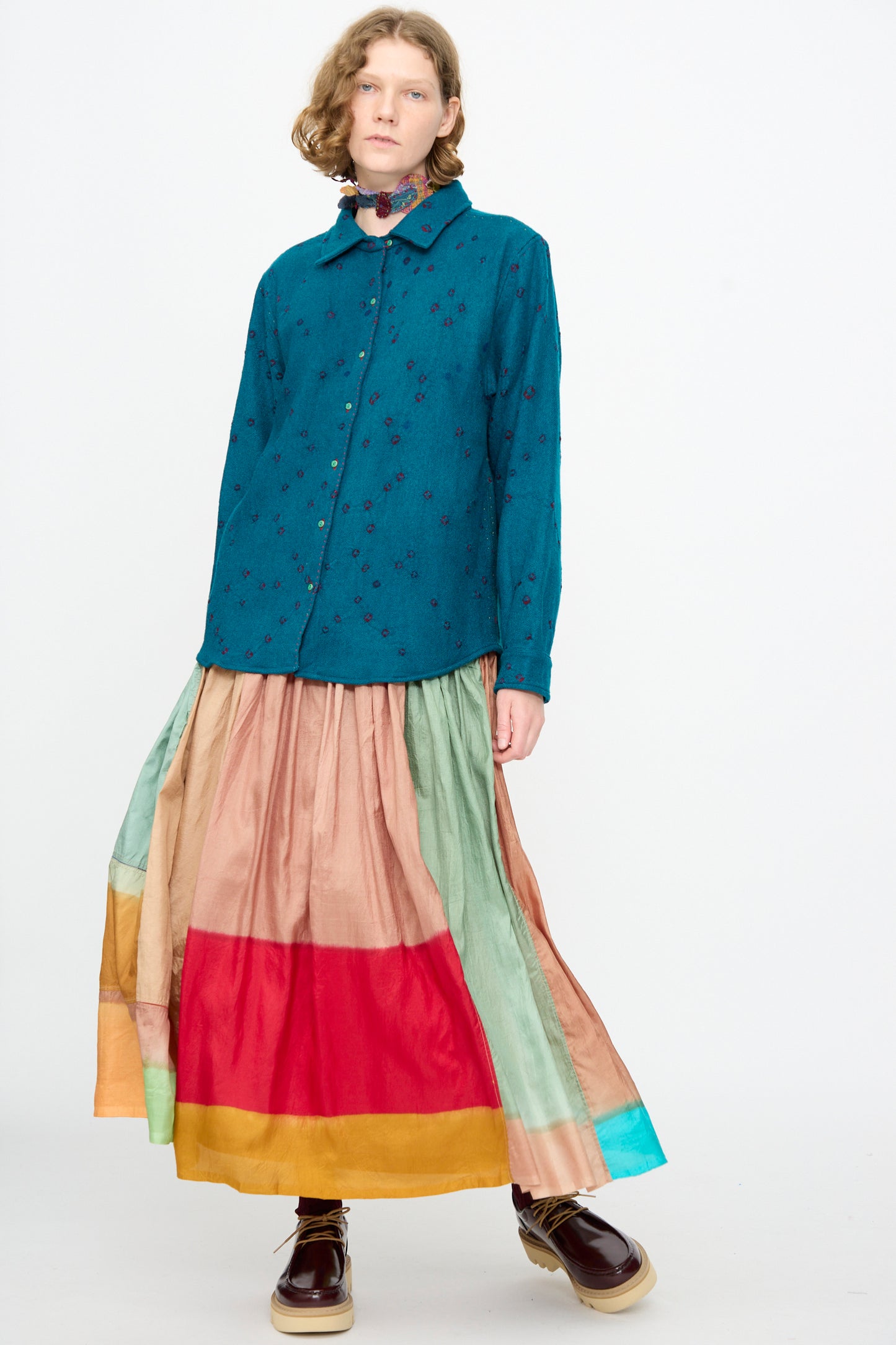 A person wearing the Injiri Bandhani Wool Shirt in Blue, featuring intricate bandhani patterns, paired with a multicolored, wide-hemmed maxi skirt and dark platform shoes—handcrafted in India—standing against a plain white background.