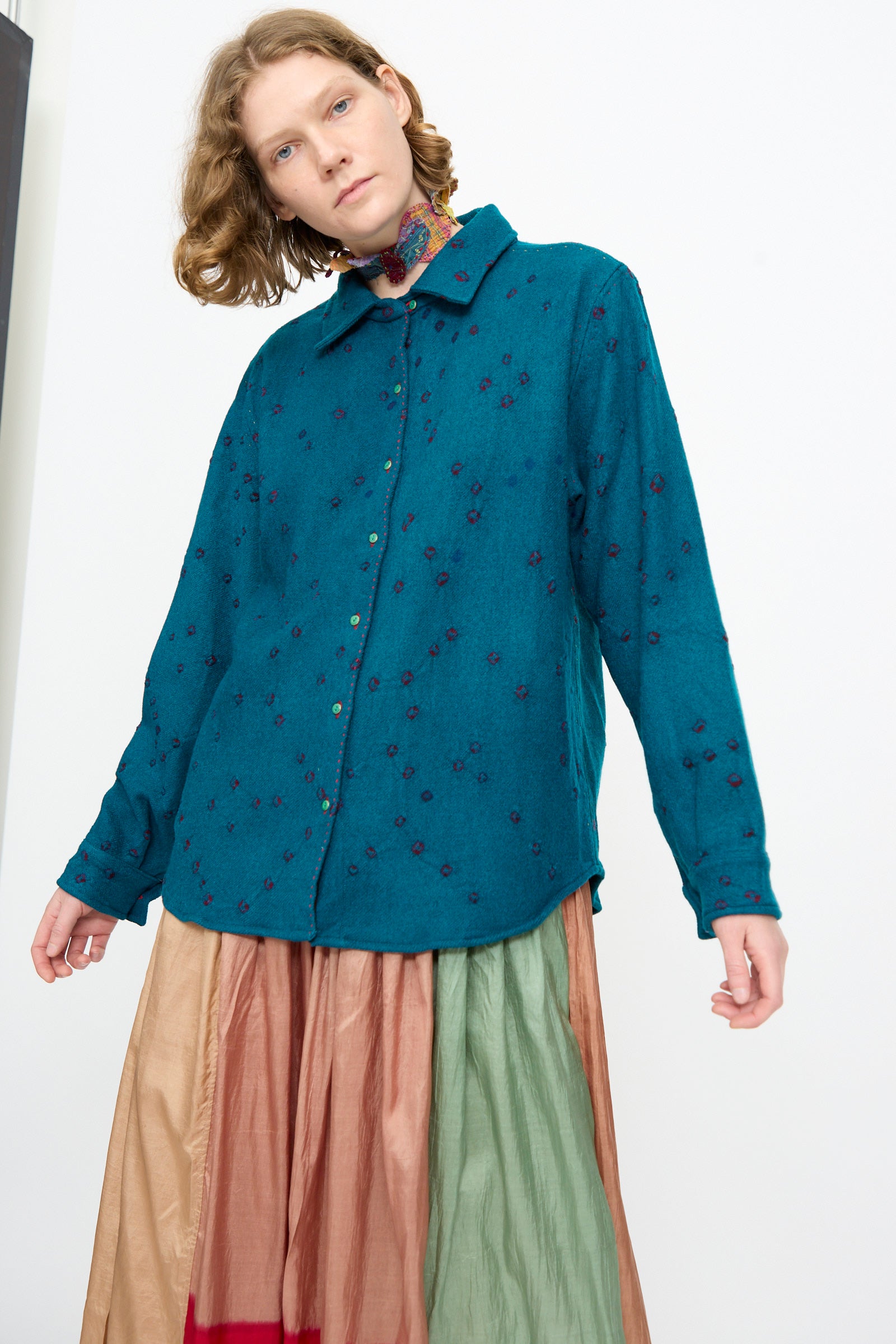 A person wearing the Injiri Bandhani Wool Shirt in Blue, handcrafted in India, along with a long multicolored skirt, stands against a plain background.