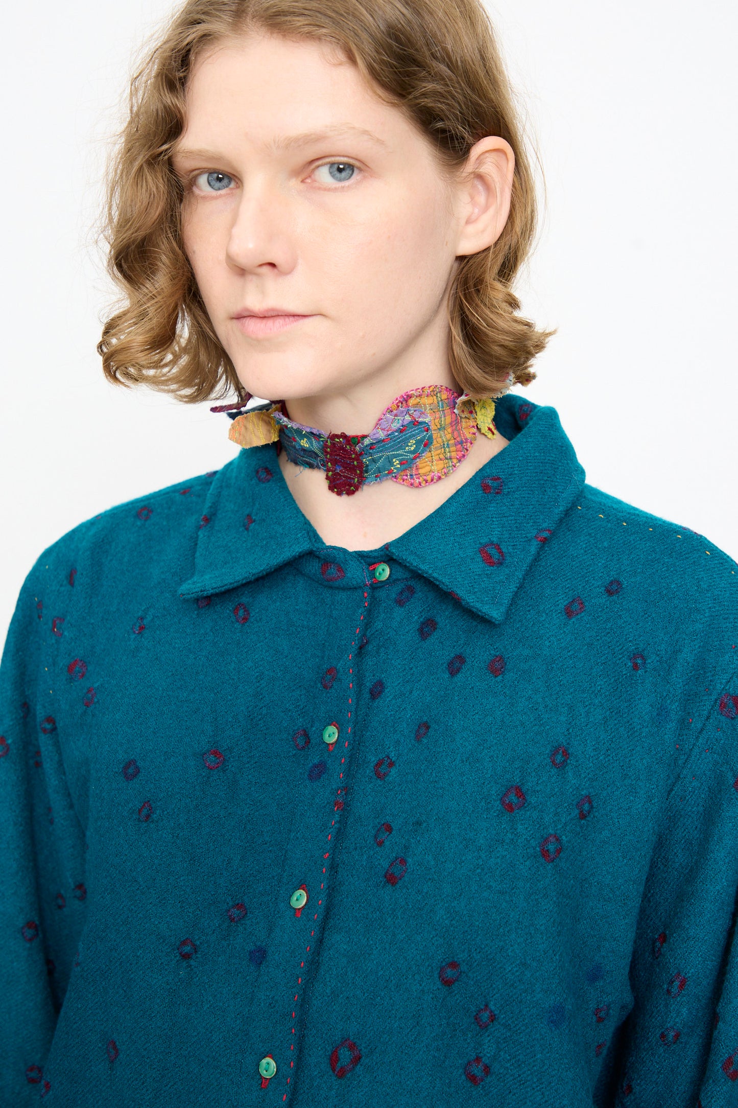 Person with short, wavy hair wearing the Injiri Bandhani Wool Shirt in Blue and a colorful, patterned fabric choker against a plain background.