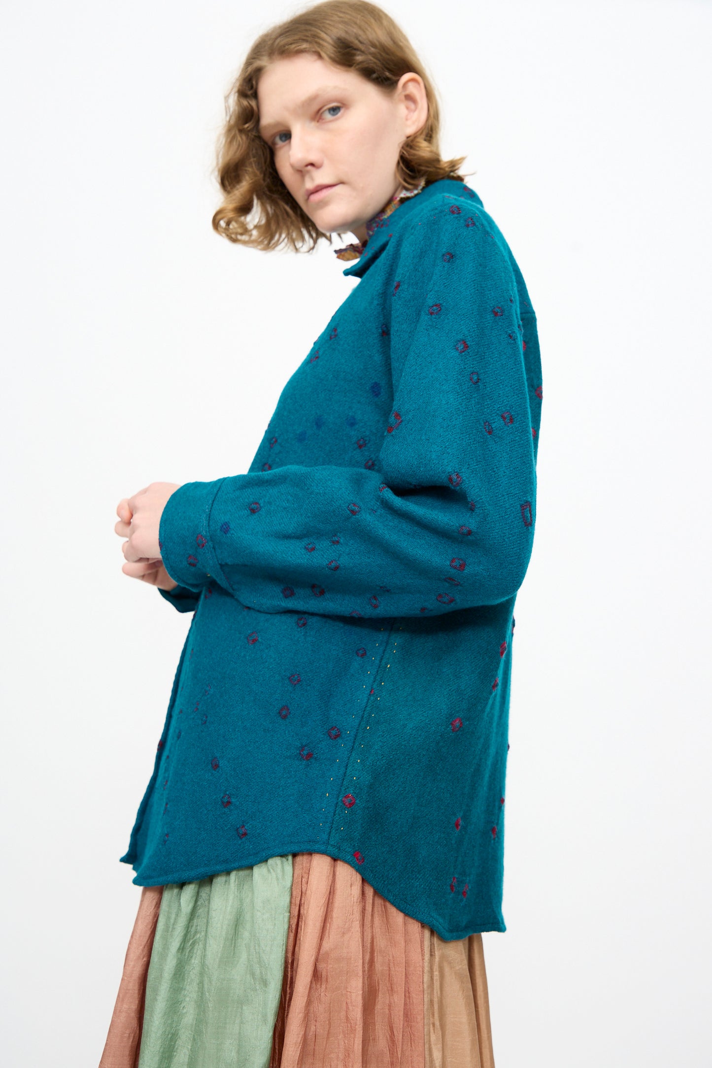 Person with short, wavy hair wearing the Injiri Bandhani Wool Shirt in Blue—handcrafted in India—paired with a pleated, multicolored skirt, standing against a plain white background.