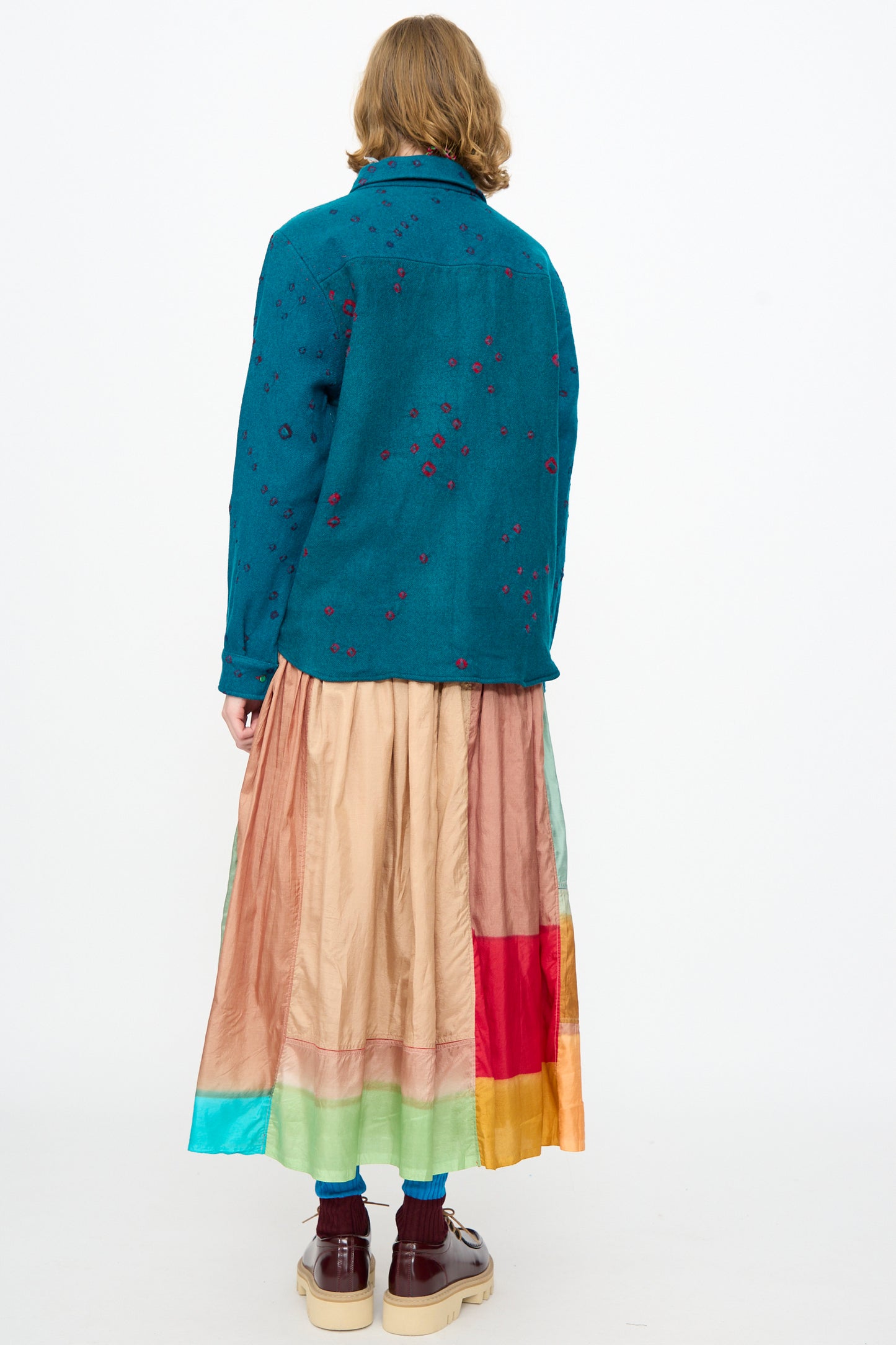 Person standing with back facing the camera, wearing a teal jacket with red details, a multicolored patchwork skirt, maroon socks, and beige shoes against a plain white background; the ensemble is thoughtfully complemented by the Injiri Bandhani Wool Shirt in Blue, handcrafted in India.
