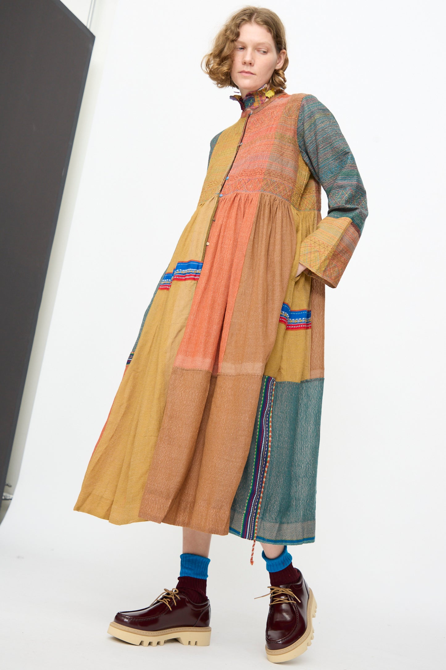 A person wearing the Bhujodi Wool and Silk Boro Jacket in Multi by Injiri, styled with blue socks and chunky brown platform shoes, standing against a plain white background.