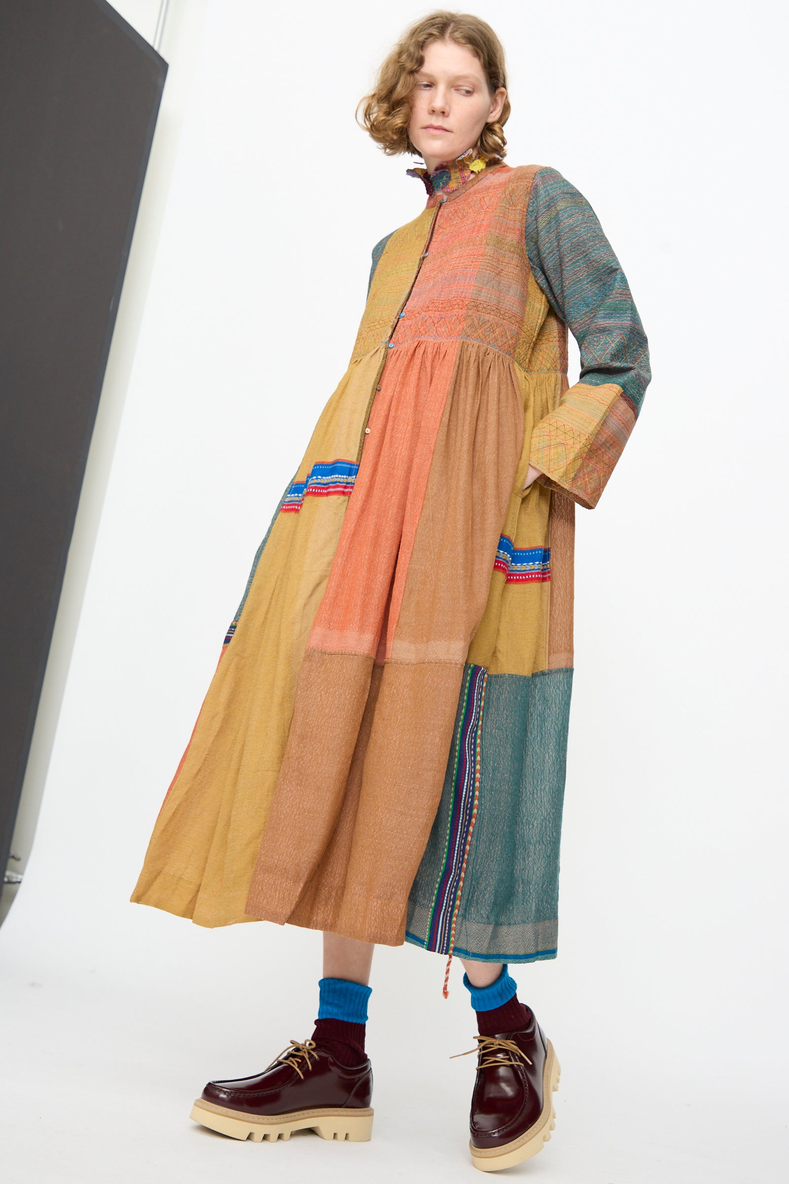 A person wearing the Bhujodi Wool and Silk Boro Jacket in Multi by Injiri, styled with blue socks and chunky brown platform shoes, standing against a plain white background.